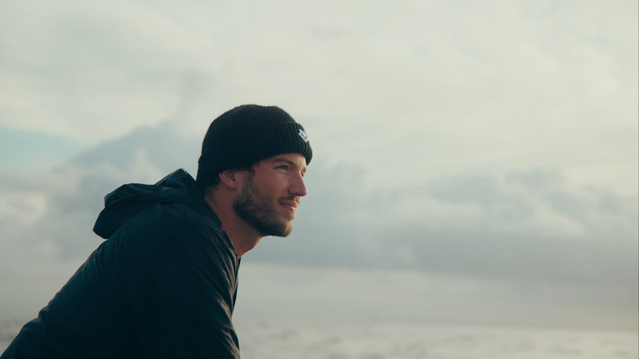 A man with a beard wearing a black beanie and jacket, looking pensively at the horizon, with a cloudy sky in the background.