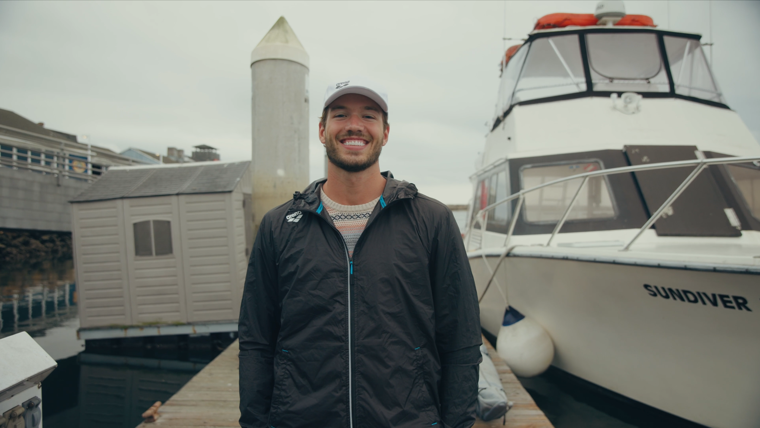 A smiling man stands on a dock near a large white yacht named 'Sundiver,' with a small gray boathouse and a lighthouse in the background.