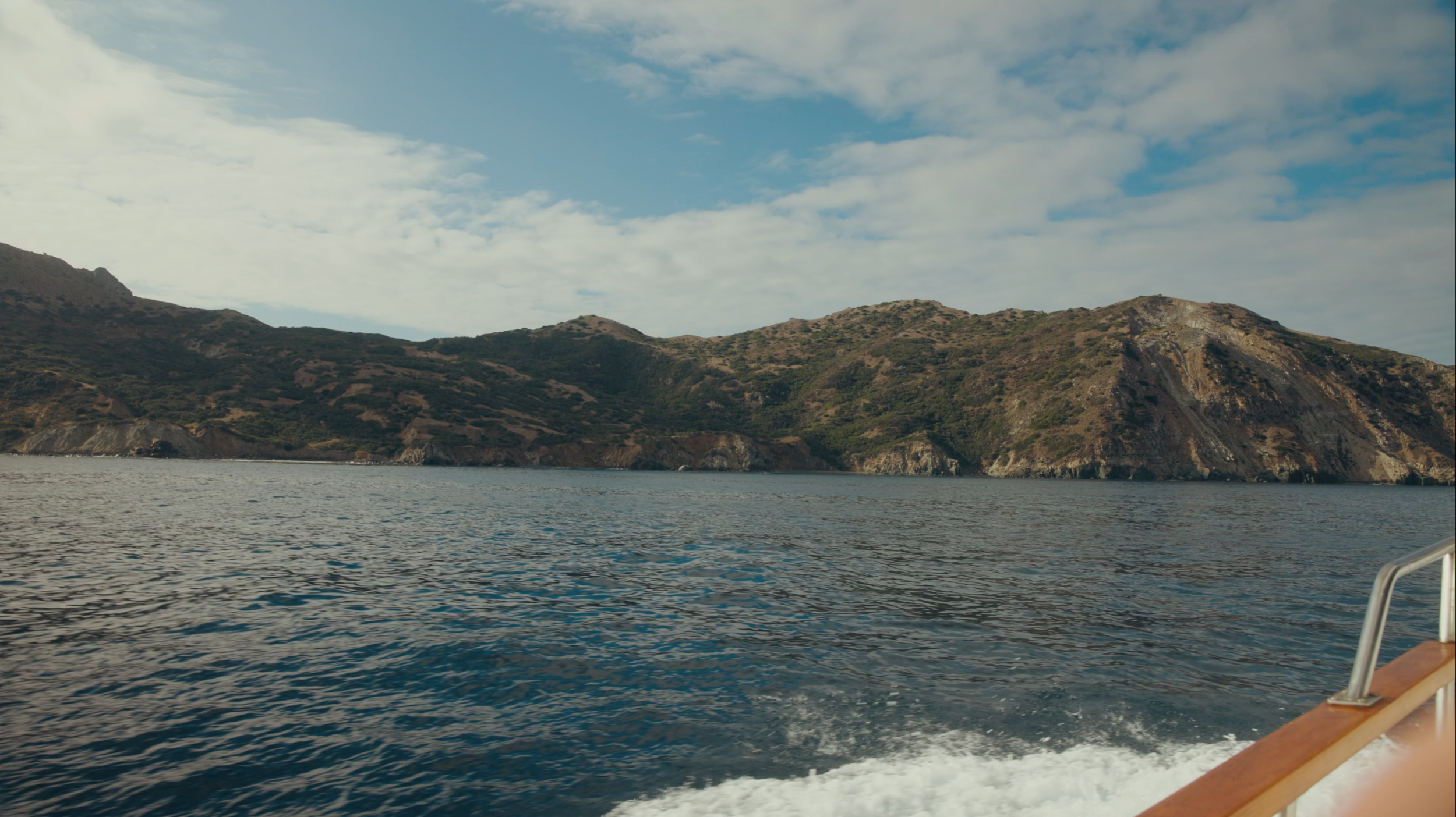 View of a hilly coastline from a boat on the water, with blue sky and clouds overhead.