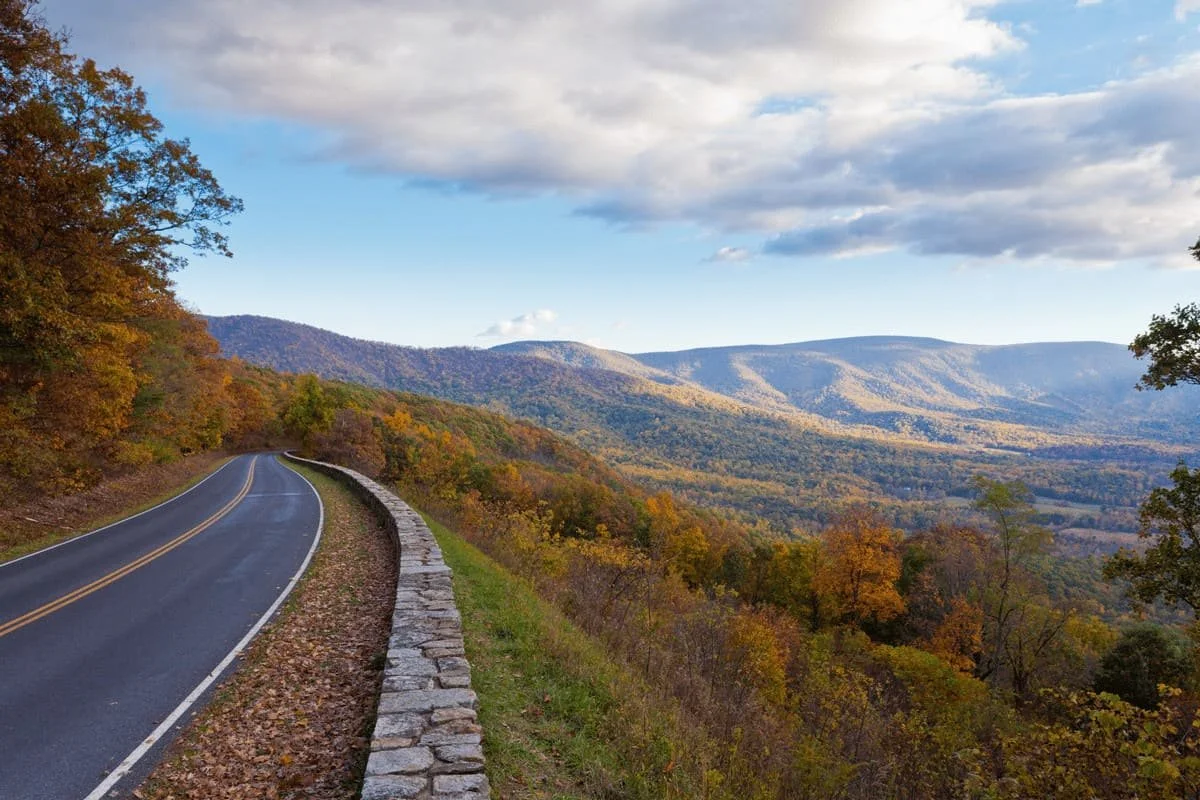 A winding mountain road with a stone wall on the right, surrounded by autumn-colored trees and distant mountains under a partly cloudy sky.