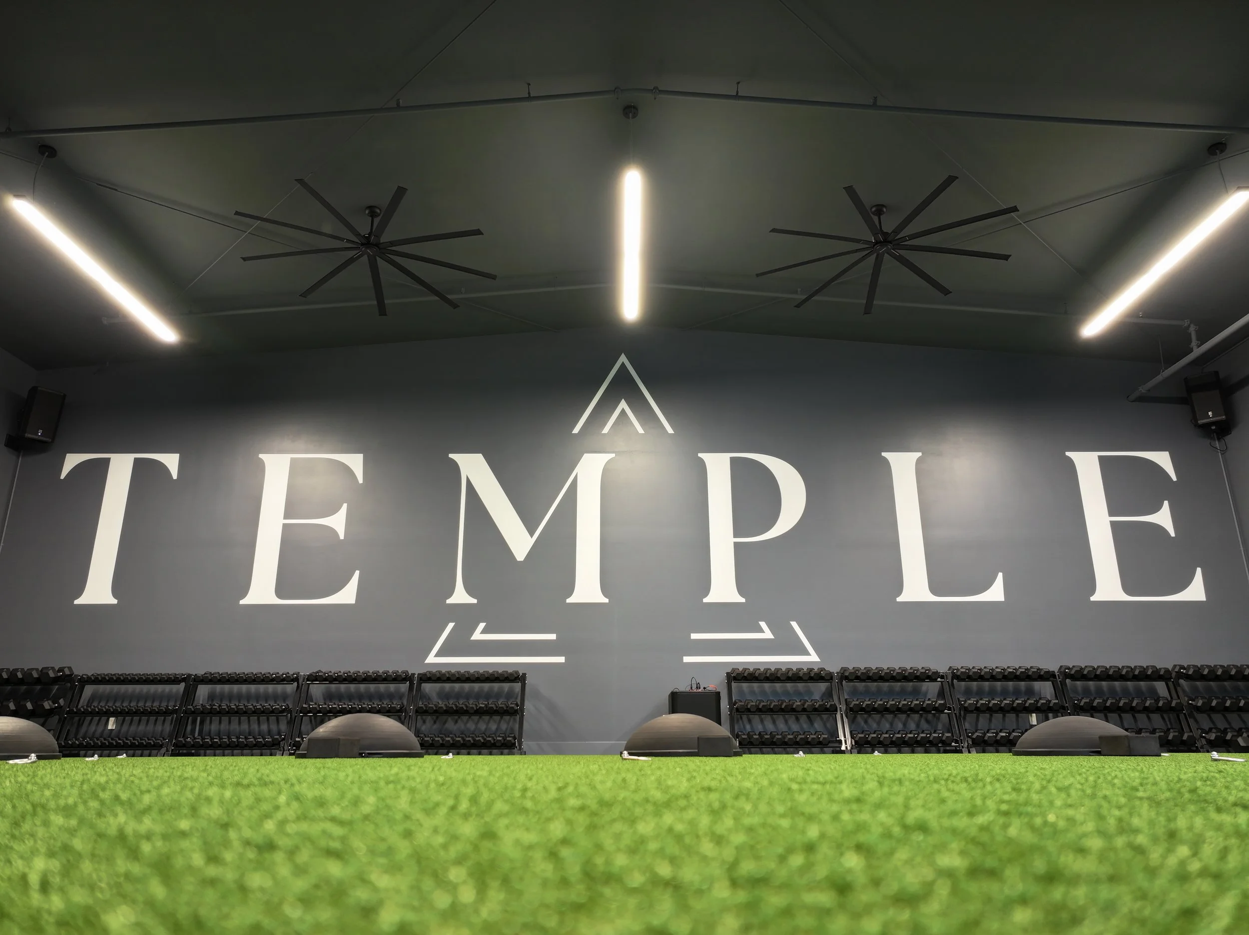 Bellingham's newest Indoor fitness gym with the word 'TEMPLE' written on the wall in large white letters, green artificial turf flooring, black workout equipment, and modern ceiling fans. Temple Fitness NW, a Pilates-style group fitness class.