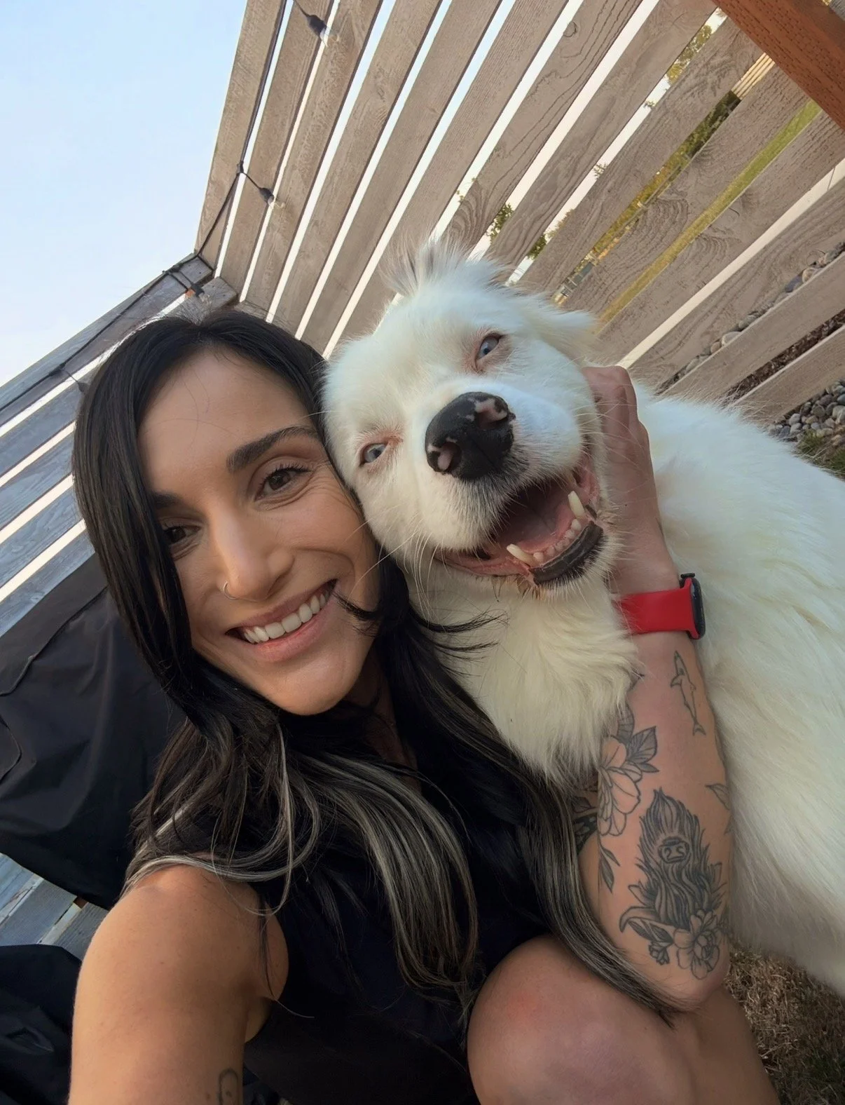 Stormy, a Temple Fitness Trainer at Bellingham, WA. She with long dark hair and tattoos is smiling while taking a selfie with a happy, white dog with one blue eye and one brown eye, outdoors near a wooden fence.