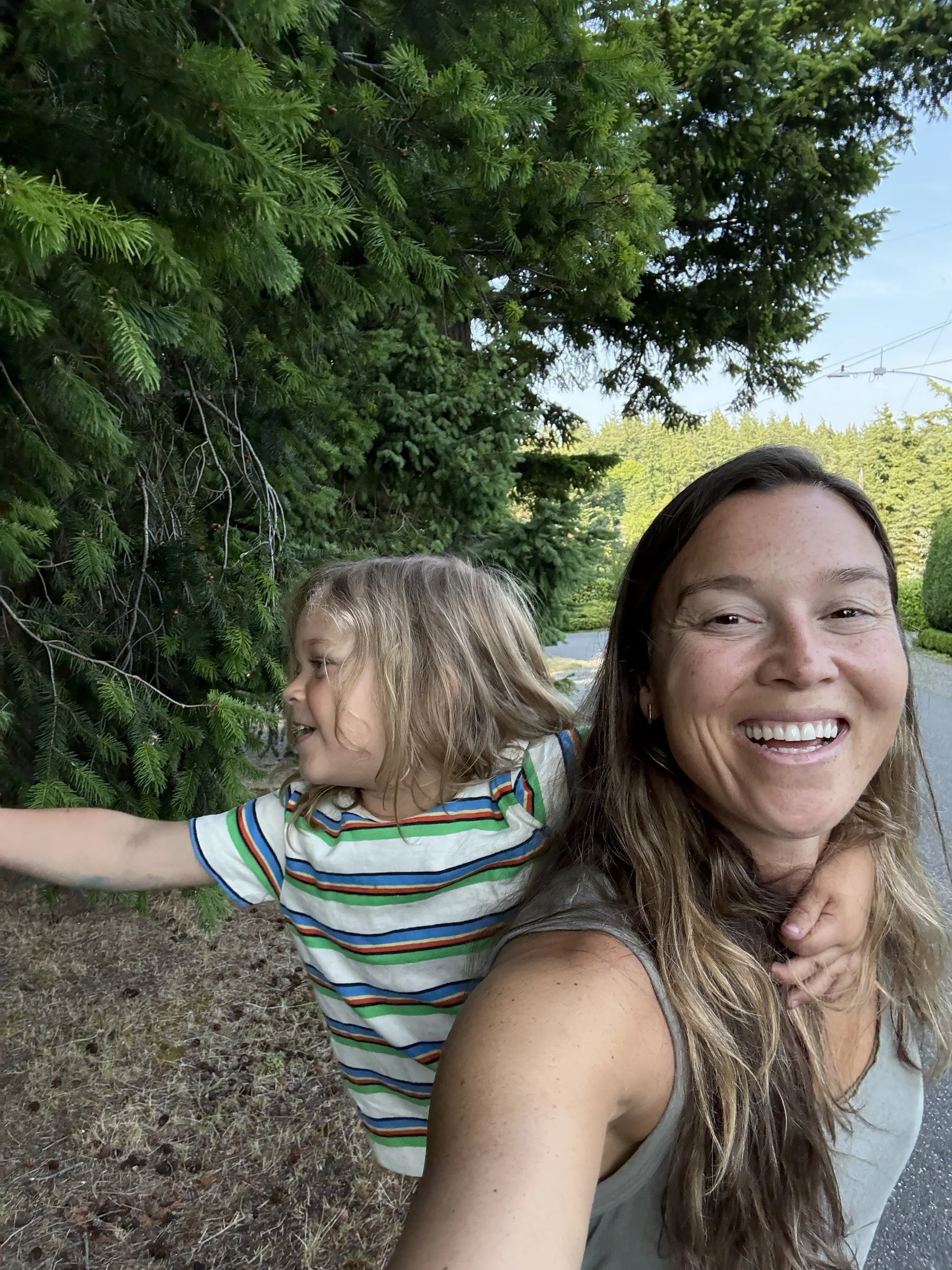 Danielle, Owner of Temple Fitness is smiling while carrying her child on her back, standing outdoors near a large evergreen tree. The child reaches out and touches the tree with their arm extended.