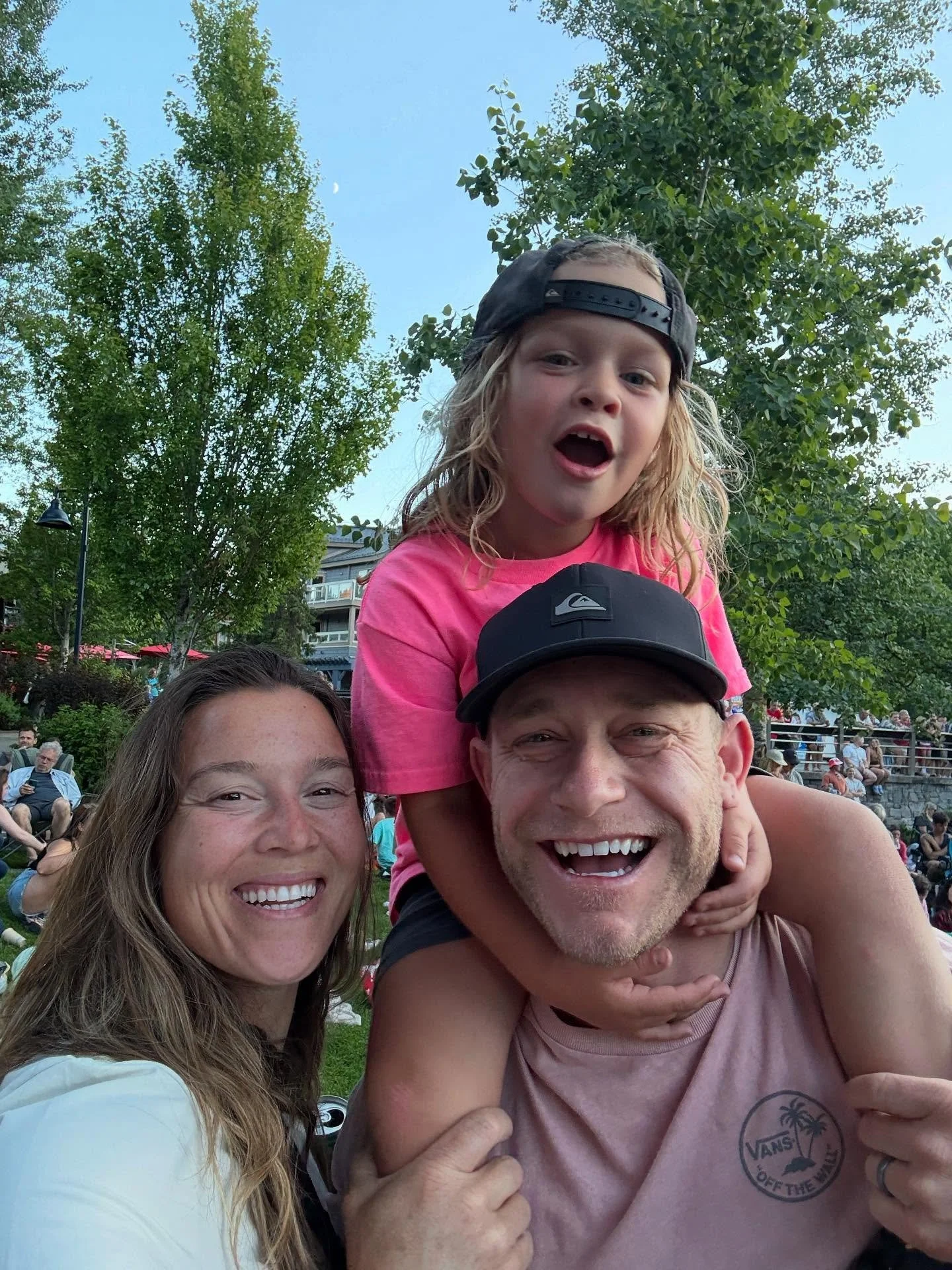 A happy family enjoying an outdoor event, with a woman, a man, and a young girl on the man's shoulders, all smiling at the camera. The background shows trees and an evening sky with moon and some people sitting on grass.