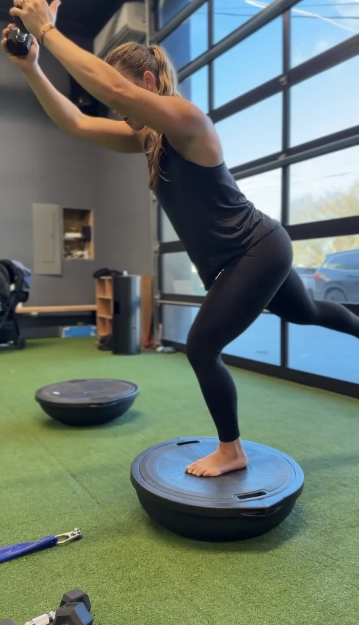 A woman exercising on a BOSU ball at the gym, using resistance bands for strength training, with large windows in the background.