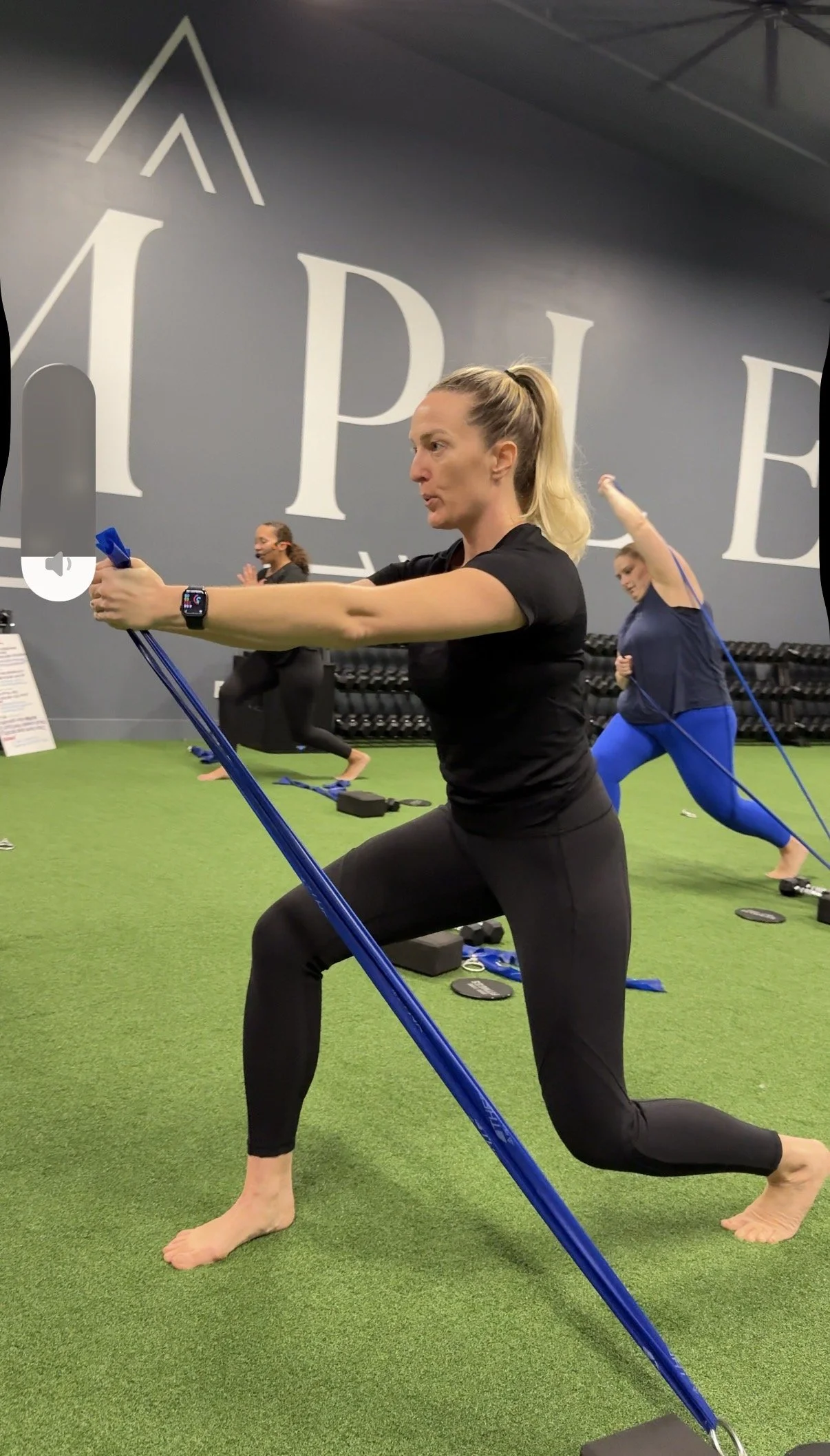 A woman doing a lunge exercise with resistance bands in a gym, with other women exercising in the background.