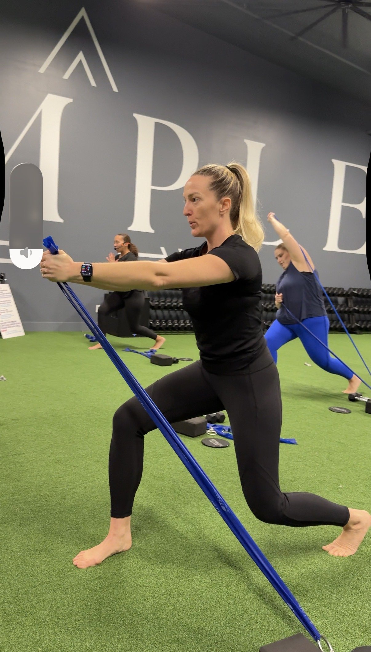 A woman doing a lunge exercise with resistance bands in a gym, with other women exercising in the background.