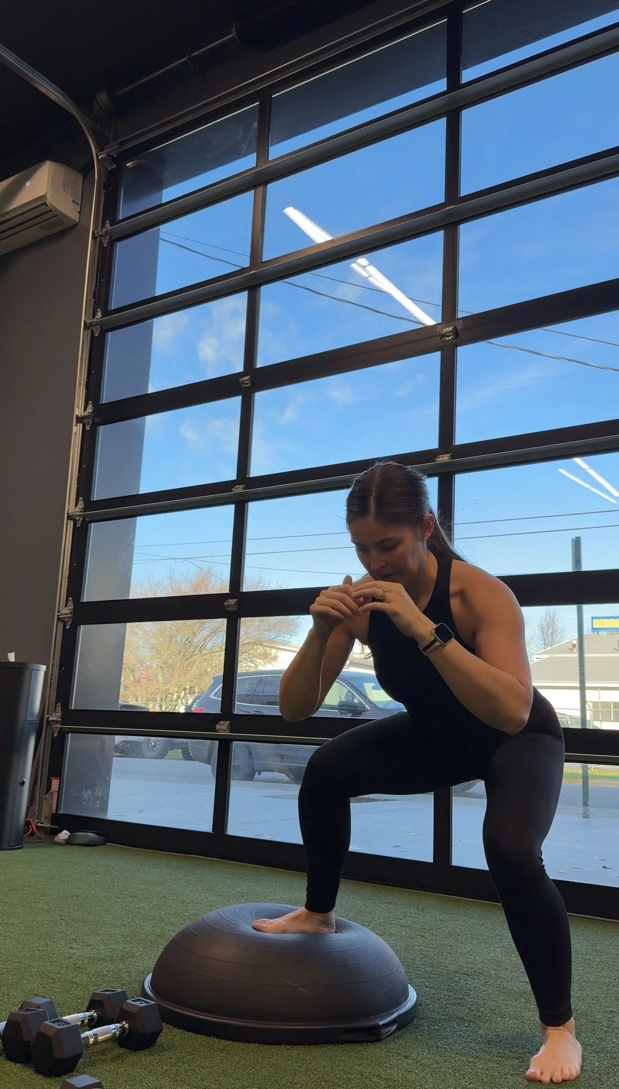 Jasmin, a Temple Trainer, in black workout clothing balances on a Bosu Ball with one foot, while holding her hands near her mouth in a gym with large garages and outside view in Bellingham, WA