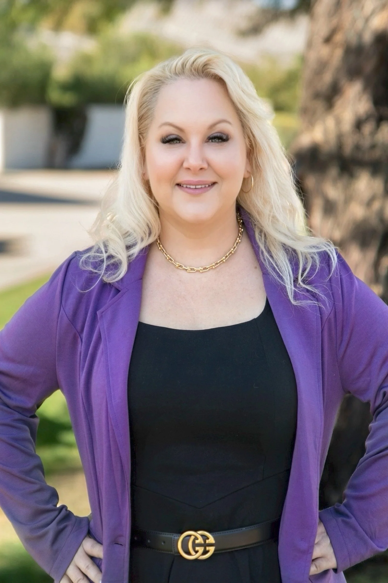 Kristie O'Brien with blonde hair, wearing a purple blazer over a black top, gold jewelry, and a black Gucci belt, standing outdoors near a tree with blurred greenery in the background.