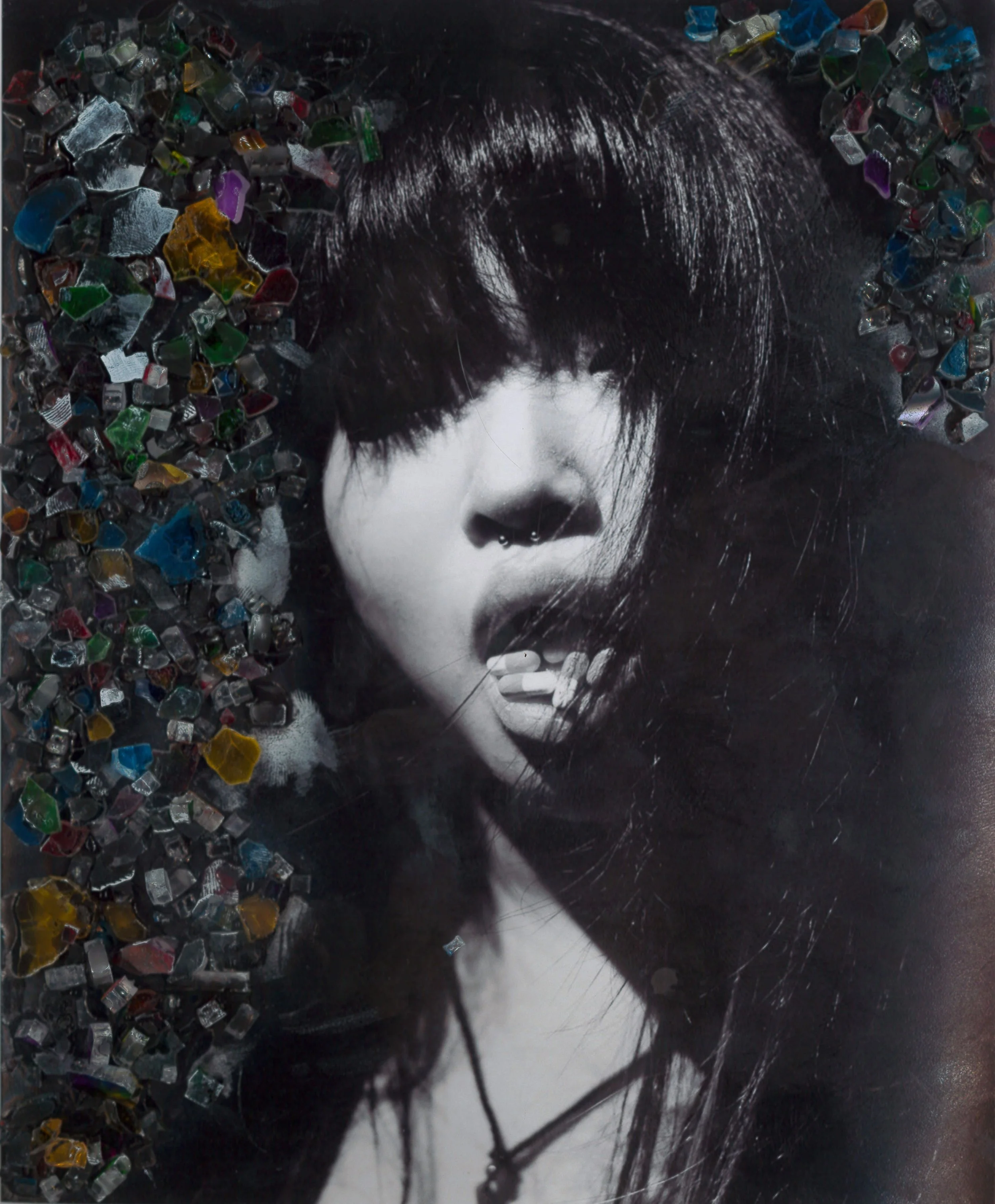 Black-and-white photograph of a woman with dark, straight hair covering one eye, exposed tongue, septum piercing, wearing a necklace, with colorful broken glass pieces on the left side of the image.