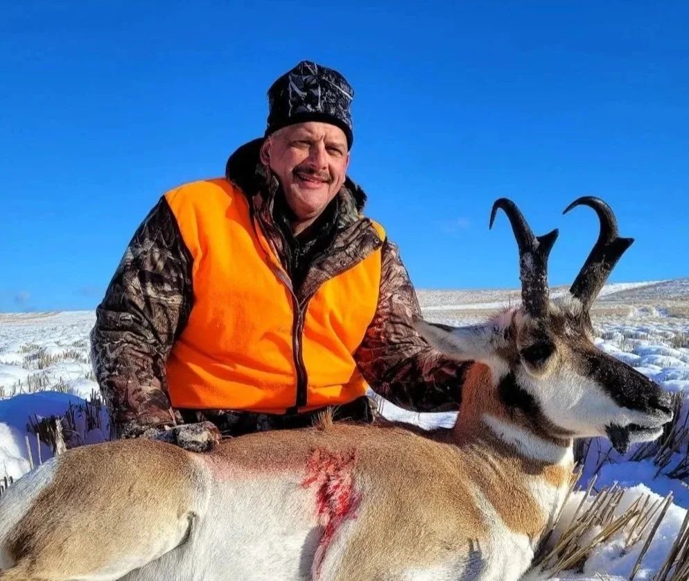 A man in camouflage and orange hunting gear kneels in snow next to a killed pronghorn antelope with two prominent horns, showing a hole and blood on its side.