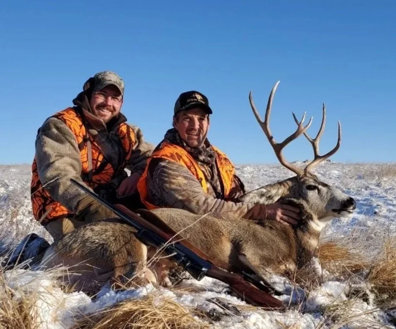 Two hunters dressed in camouflage and orange safety vests are sitting on the snow-covered ground, holding a large deer with impressive antlers.