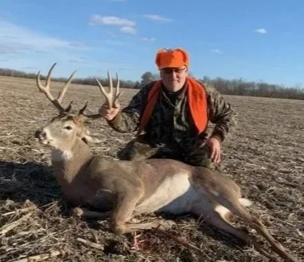 Hunter in camouflage and orange gear kneeling next to a large, freshly hunted deer with antlers, in an open field under a blue sky.