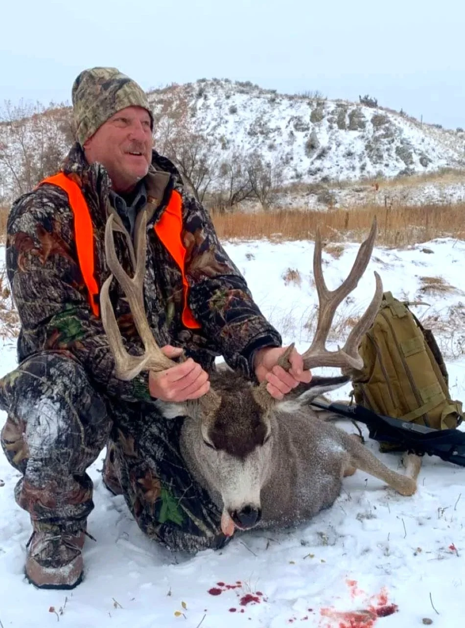 A man in camouflage hunting gear and an orange safety vest kneeling in the snow, holding the antlers of a large, dead deer with a snowy hill in the background.