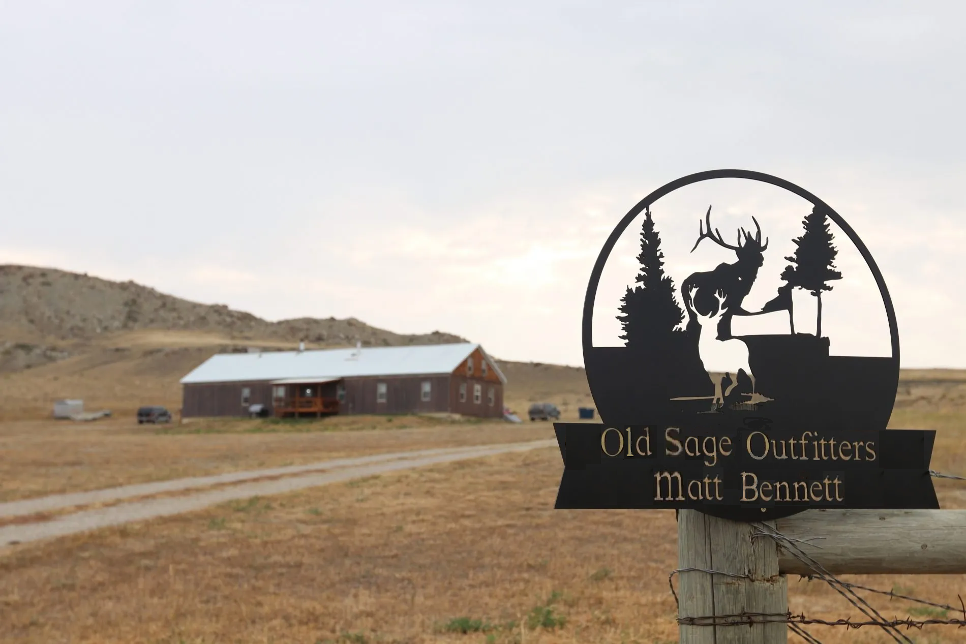 A black metal sign that reads 'Old Sage Outfitters Matt Bennett' features a silhouette of a deer with large antlers, trees, and mountains in the background. The sign is mounted on a wooden post in a rural landscape with a building, parked cars, and r