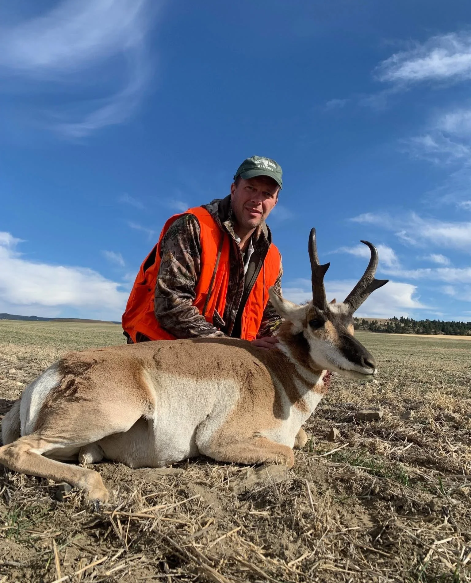 A man wearing camouflage and an orange hunting vest kneels next to a pronghorn antelope he has hunted, lying on the ground in an open field under a blue sky with some clouds.