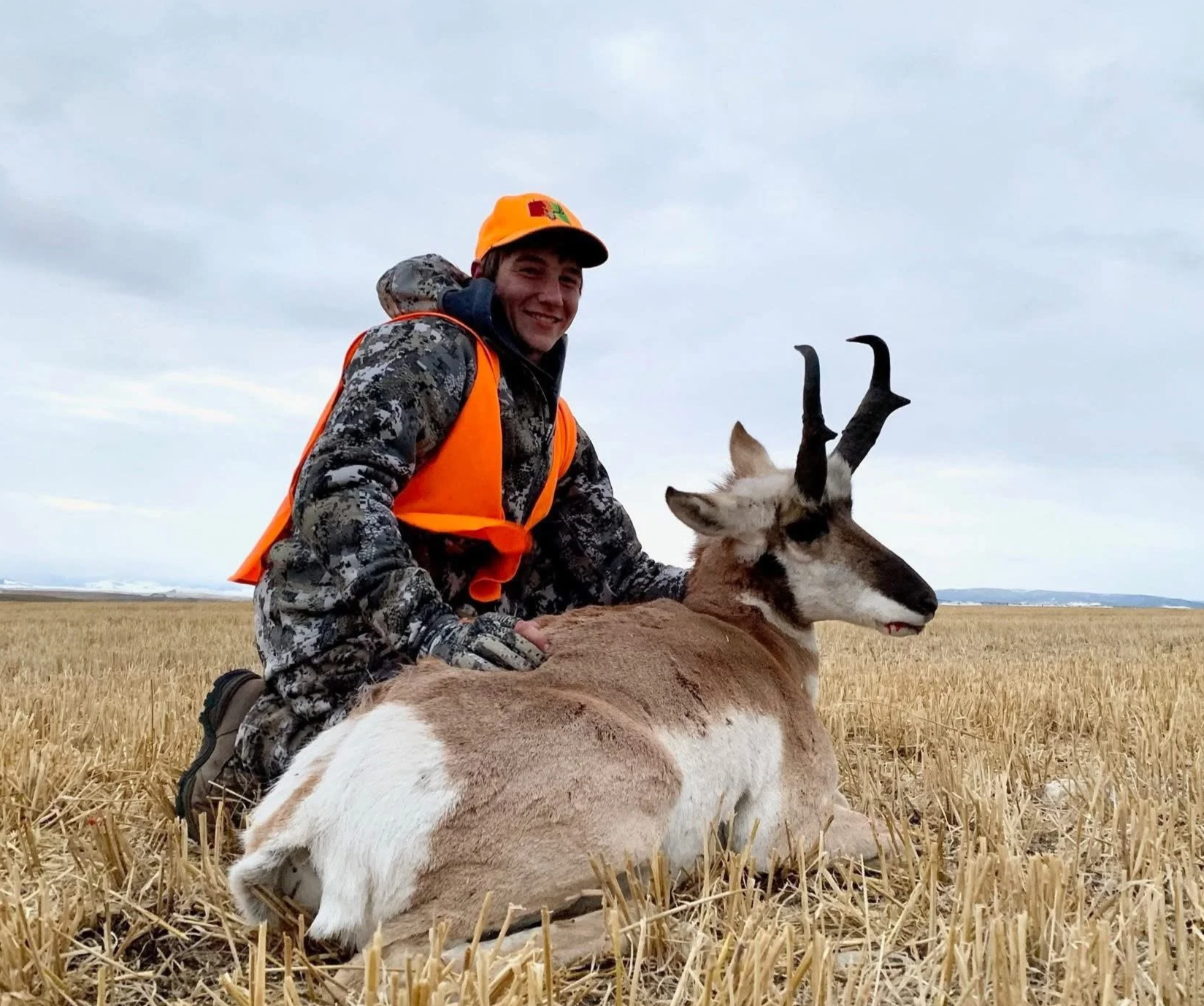 A young man in camouflage clothing, orange hunting vest, and orange hat kneels beside a resting antelope with twisted horns in a harvested field under cloudy sky.