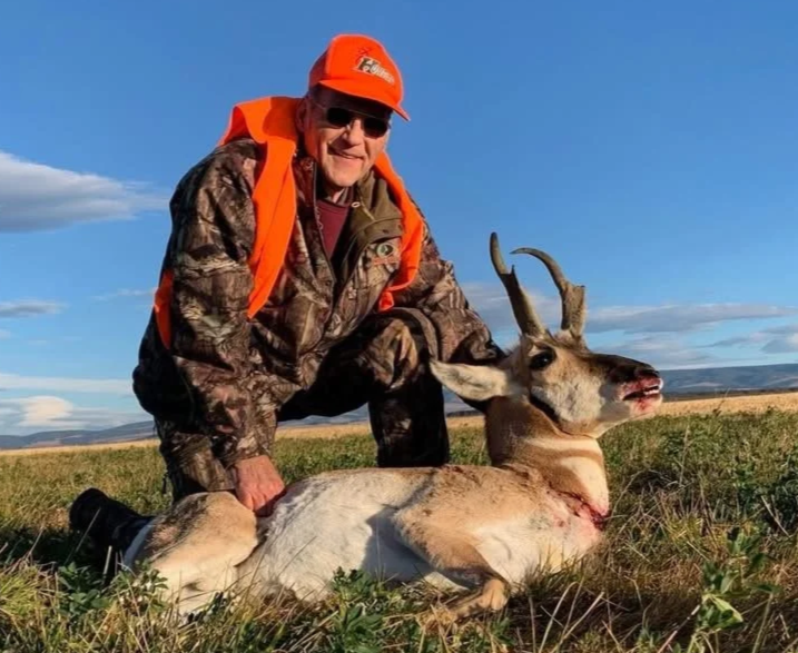 Older man in camouflage clothing and orange hat kneeling next to a freshly hunted antelope lying on the ground. The man is smiling, wearing sunglasses, in an open field with a blue sky and some clouds.