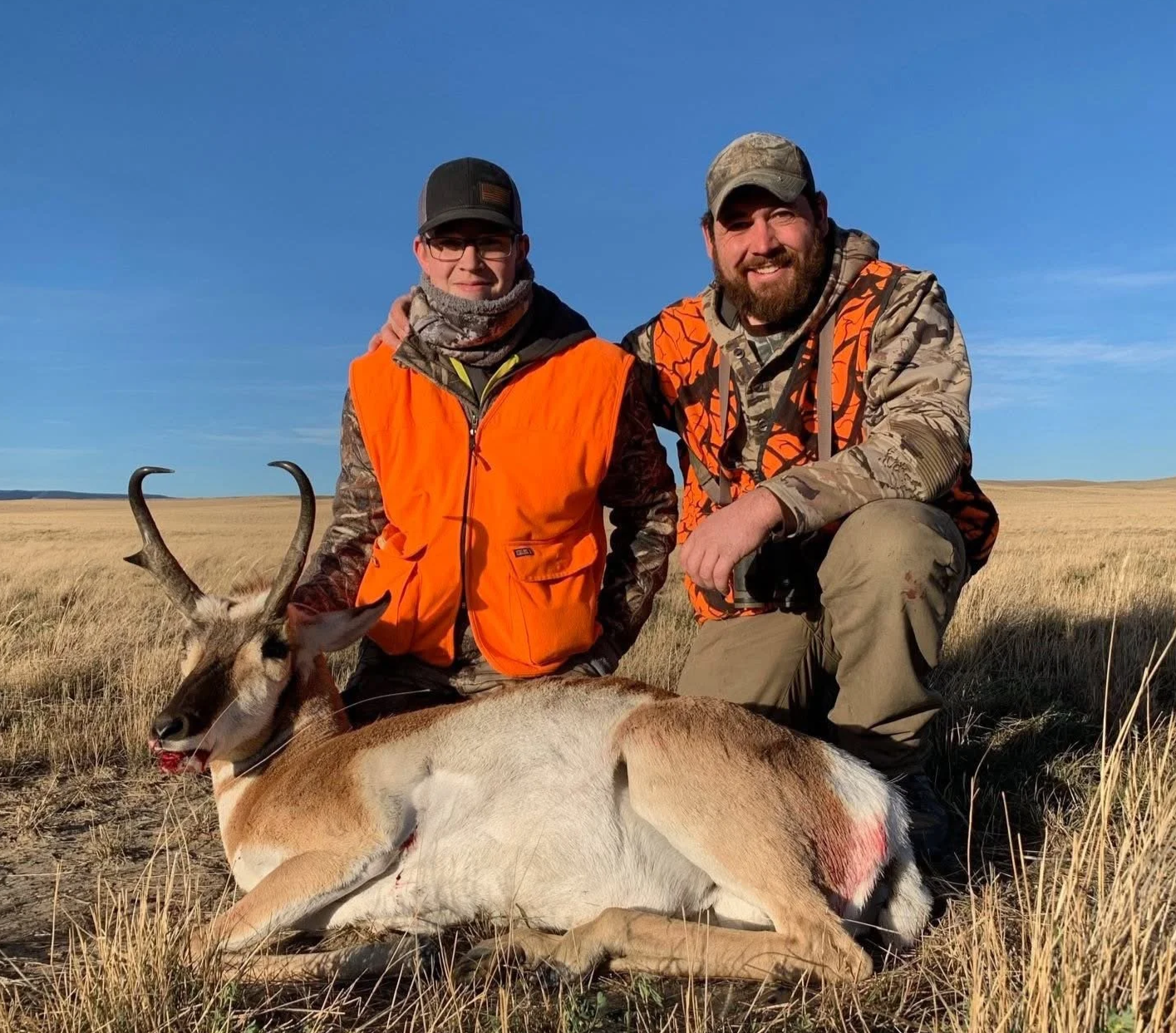 Two hunters in camouflage and orange vests kneel in a grassy field with a fallen pronghorn antelope, under a clear blue sky.
