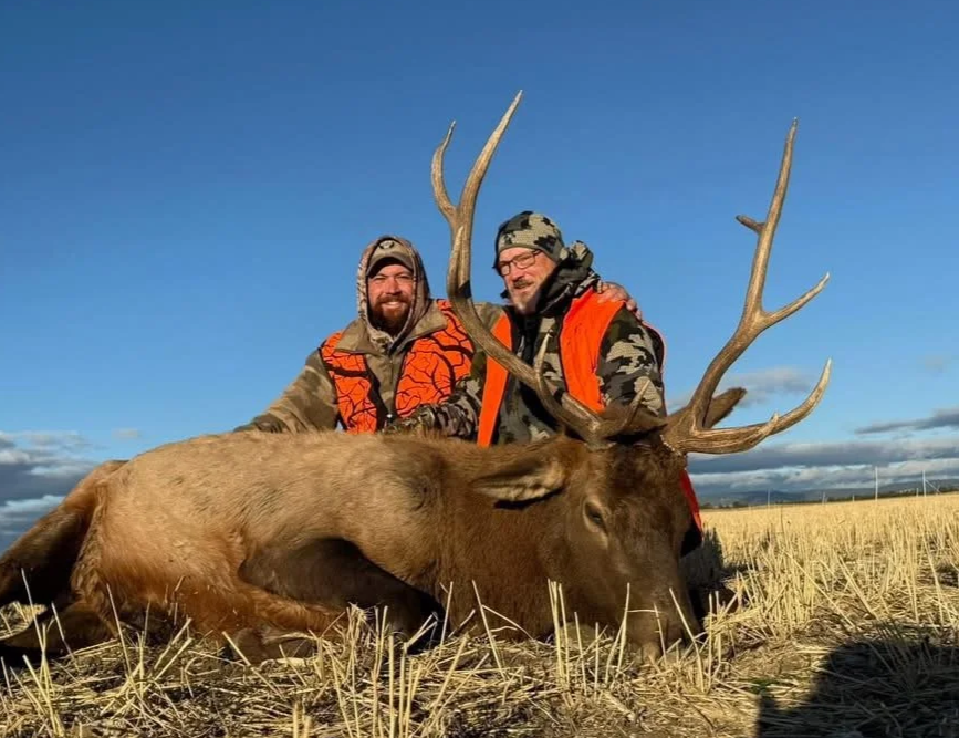 Two hunters in camouflage and orange hunting vests kneel behind a large, slain elk with impressive antlers in an open field under a blue sky.
