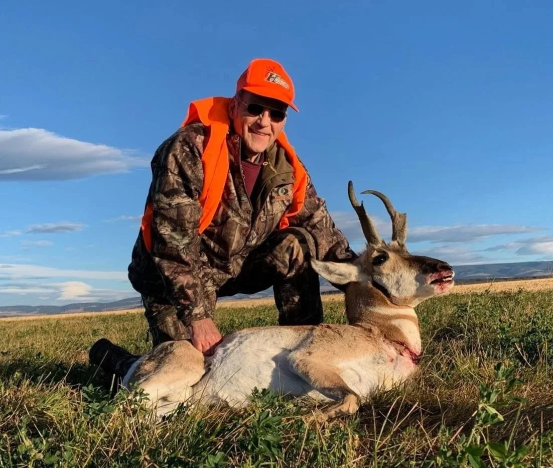 A man dressed in camouflage and orange hunting gear kneels beside a freshly hunted antelope in an open field under a blue sky with scattered clouds.