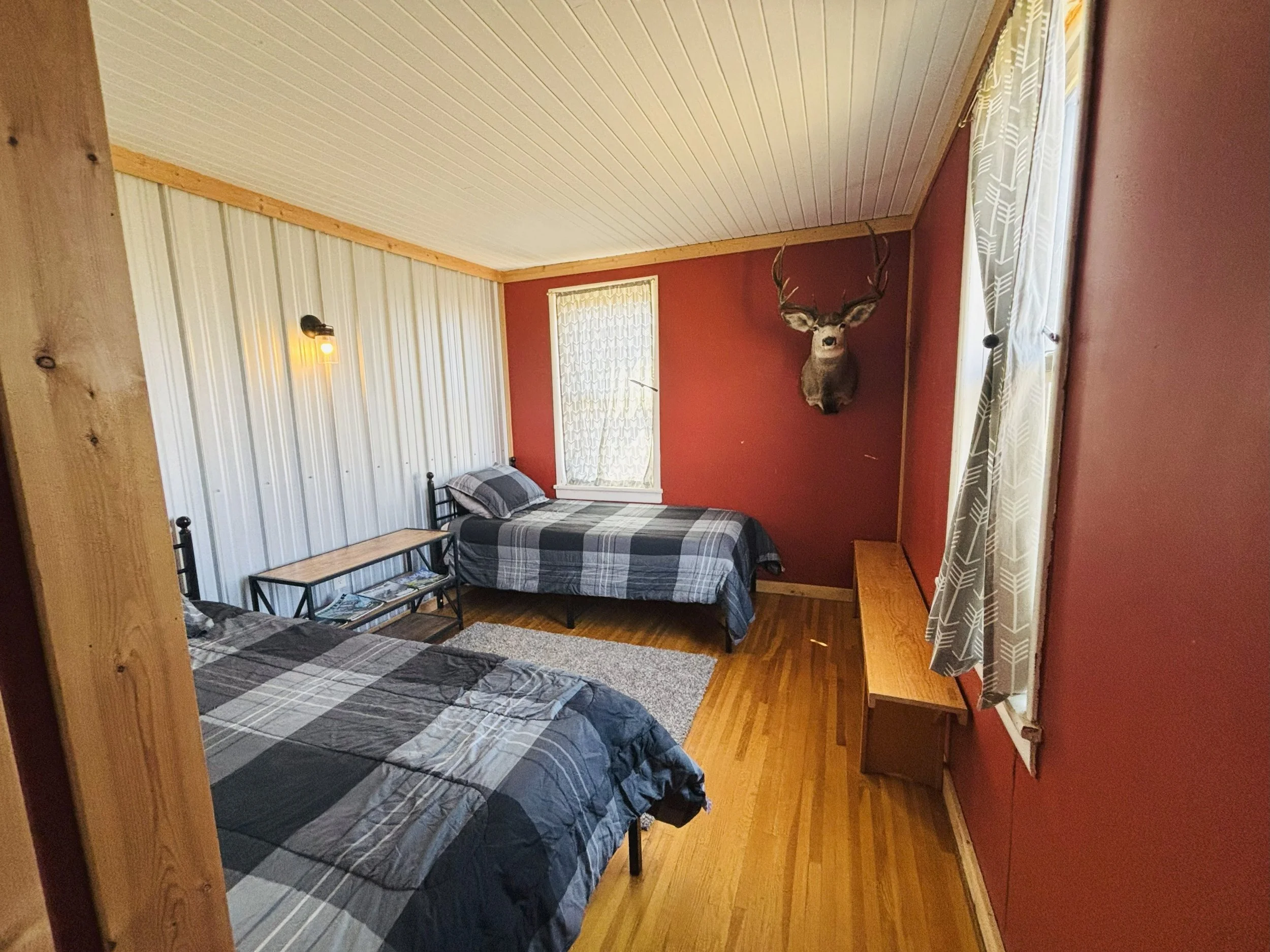 A rustic bedroom with two twin beds with plaid bedding, a wooden bench, and a mounted deer head on the back wall. The room has red and white paneled walls, hardwood floors, and windows with patterned curtains.