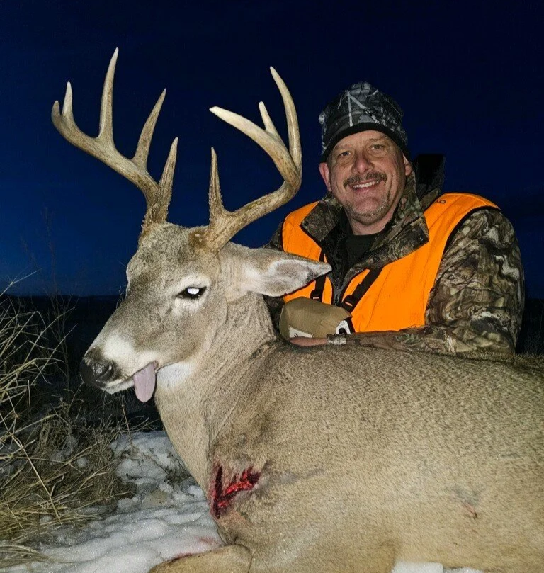 A smiling man in camouflage and orange hunting gear poses beside a large deer with antlers, lying on the snow, with a visible wound on its side.