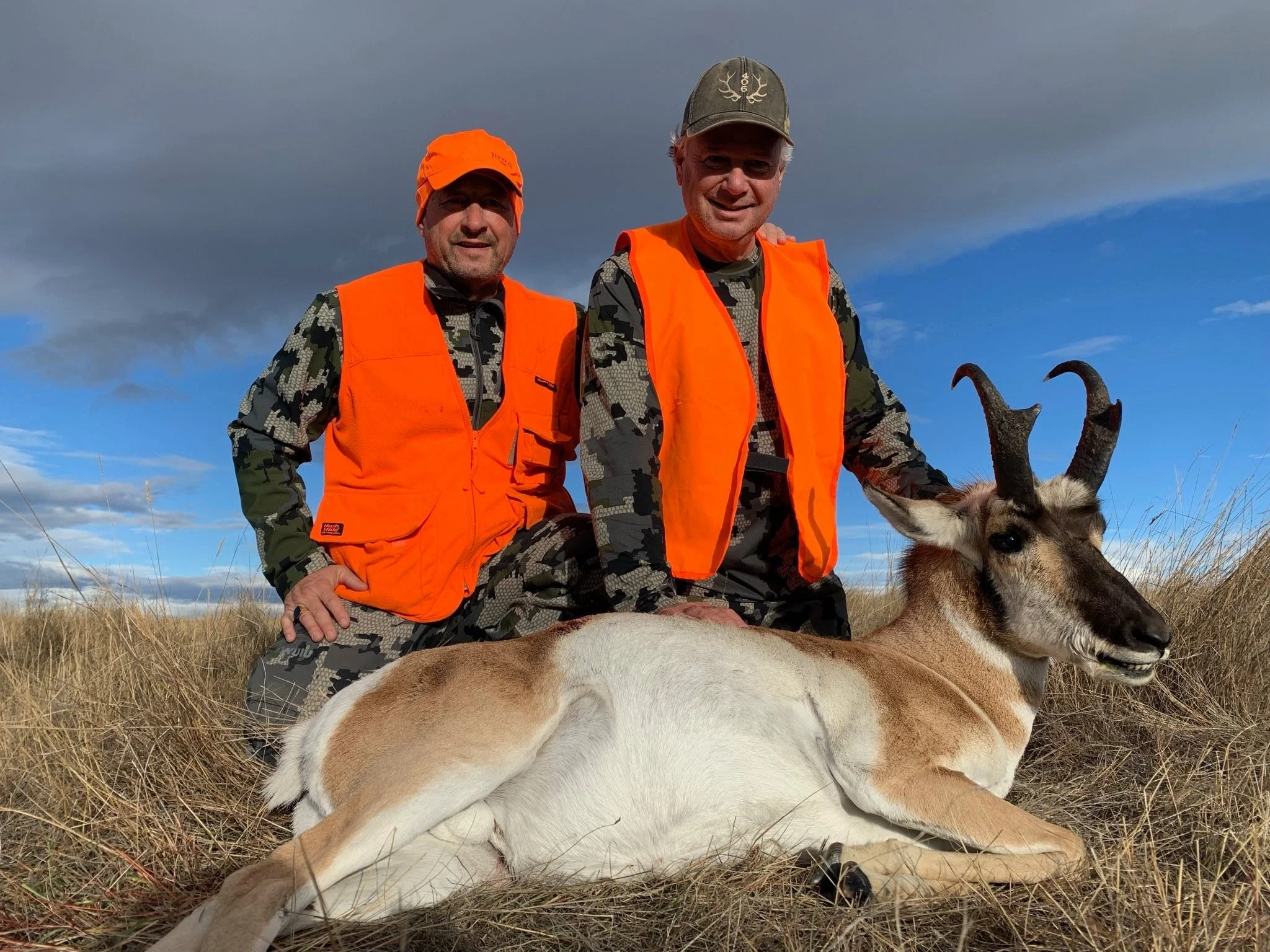 Two hunters kneeling in a grassy field during daytime, wearing orange safety vests and camouflage clothing, posing with a freshly hunted pronghorn antelope lying on the ground, with a cloudy sky in the background.