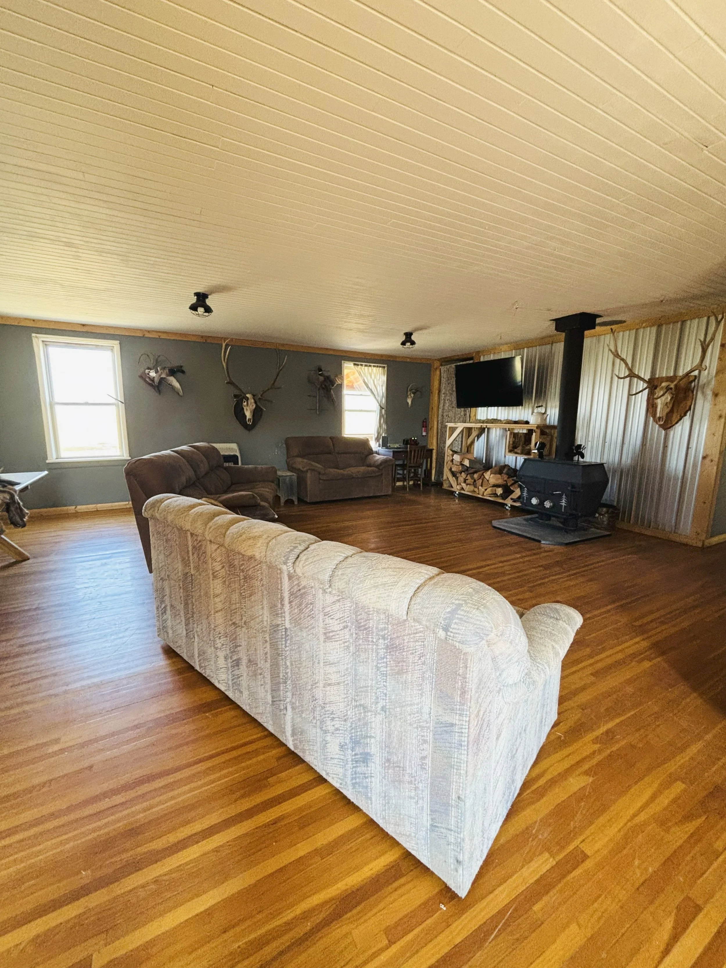 A spacious living room with hardwood floors, beige, brown, and gray sofas, hunting-themed wall decor including mounted deer skulls and antlers, a wood-burning stove, a large flat-screen TV, and windows allowing natural light.