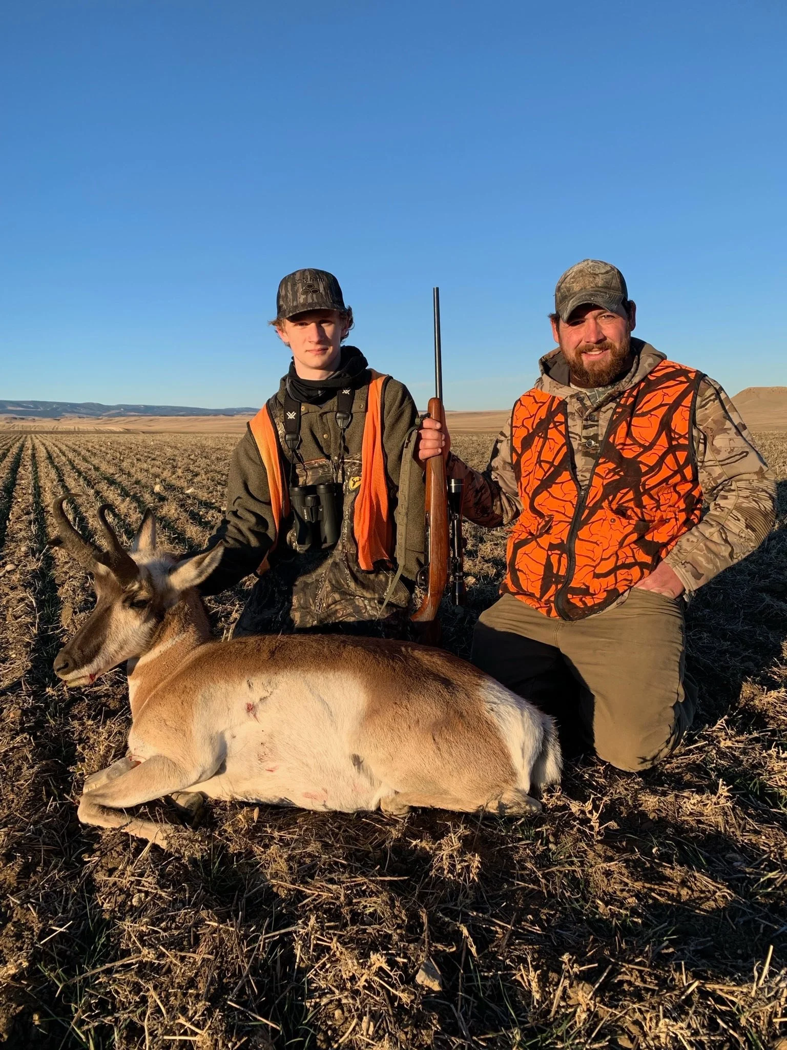 Two hunters kneeling in a field at sunset, with a freshly killed deer laying in front of them. One of the hunters is holding a rifle, and both are dressed in camouflage and hunting gear.