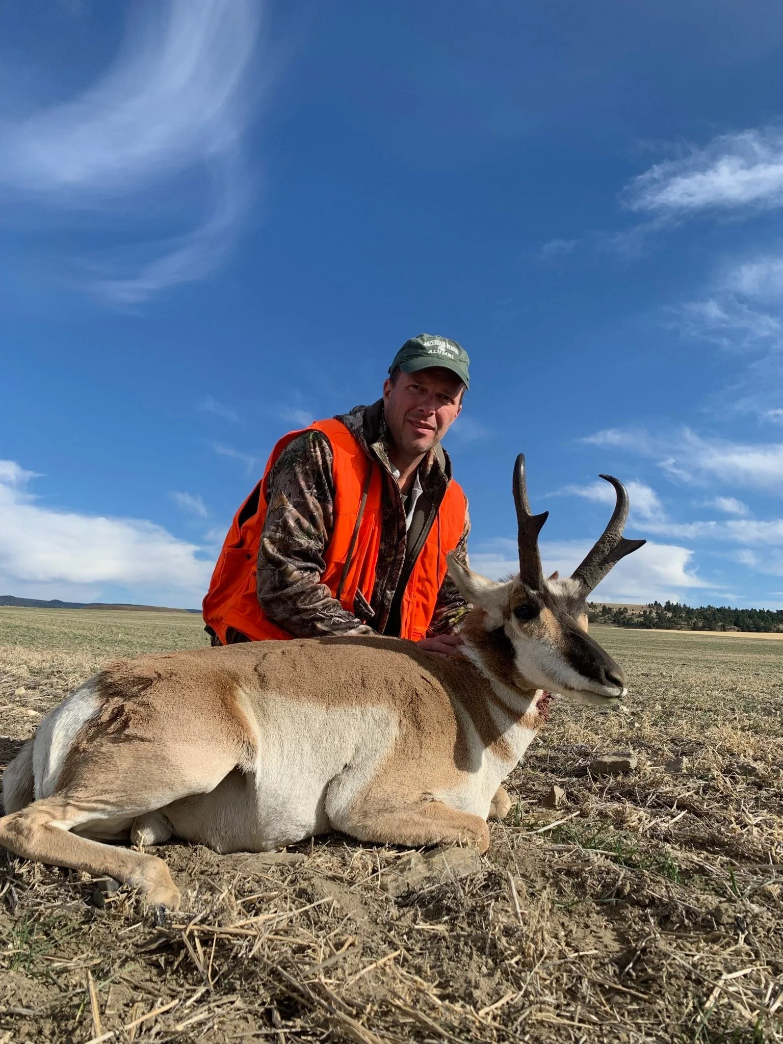 A man in camouflage and an orange vest kneels next to a lying pronghorn antelope in an open field under a blue sky with some clouds.