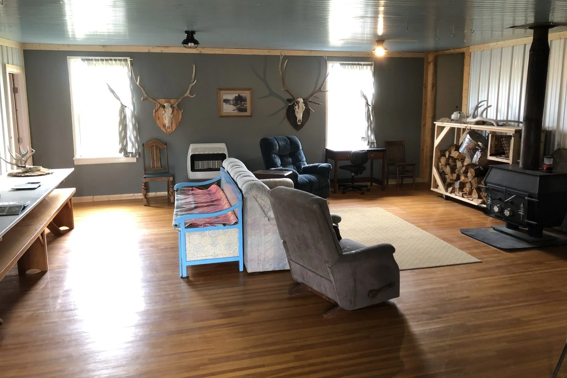 Living room with wood flooring, cluttered furniture including a recliner, a couch, a blue bench, and a wood stove, decorated with mounted animal skulls and antlers on the walls, a fireplace, and two windows with curtains.