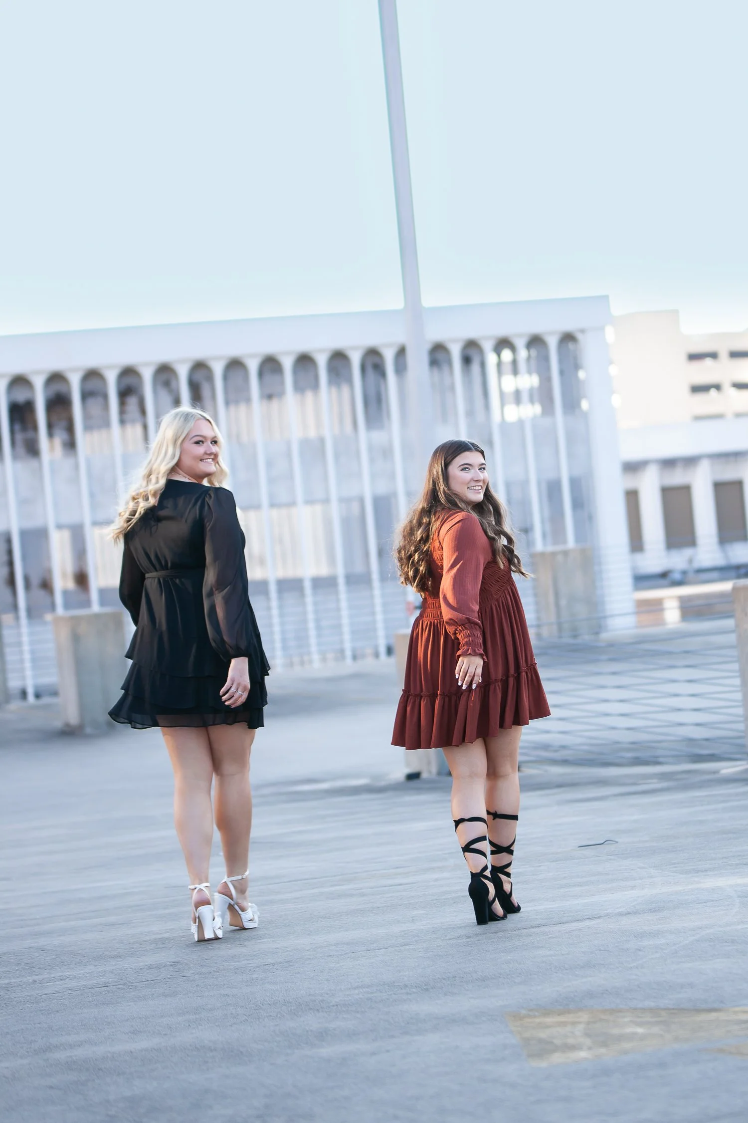 Two women in fashionable dresses and high heels walking outdoors on a rooftop.