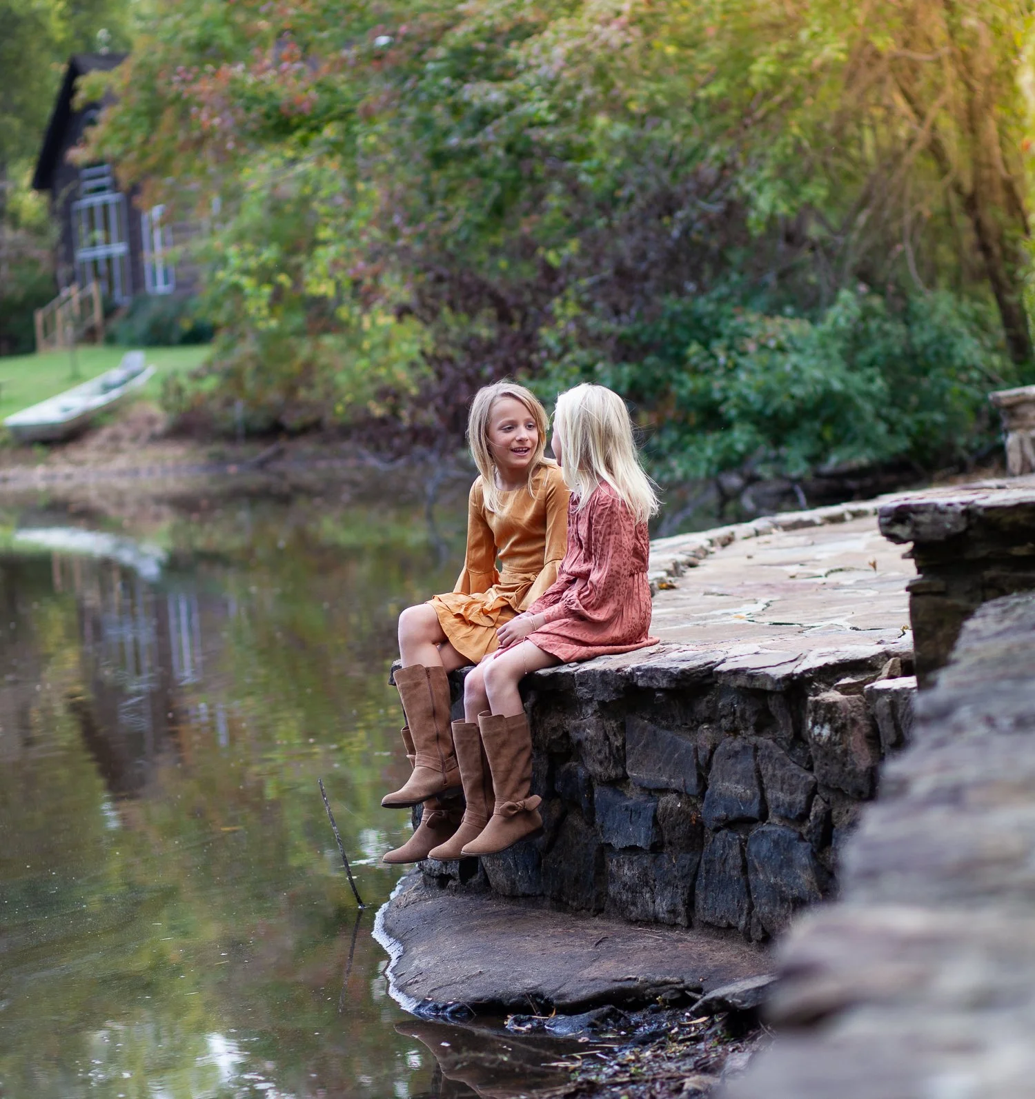 Two young girls sitting on a stone ledge by a lake, chatting, with trees and a house in the background.