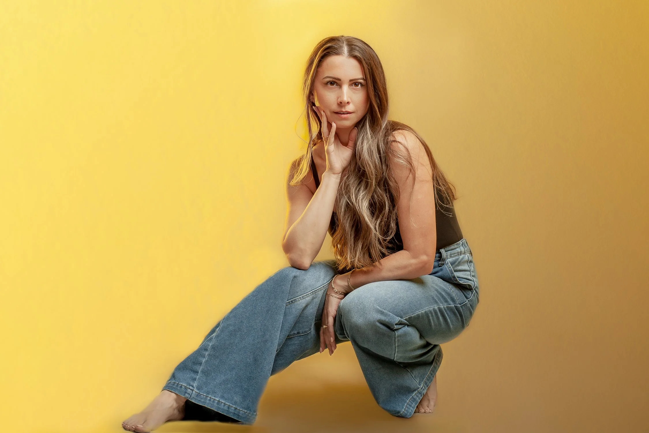 A woman with long, wavy hair dressed in a black top and blue jeans sitting against a yellow background, looking into the camera with her hand near her face.