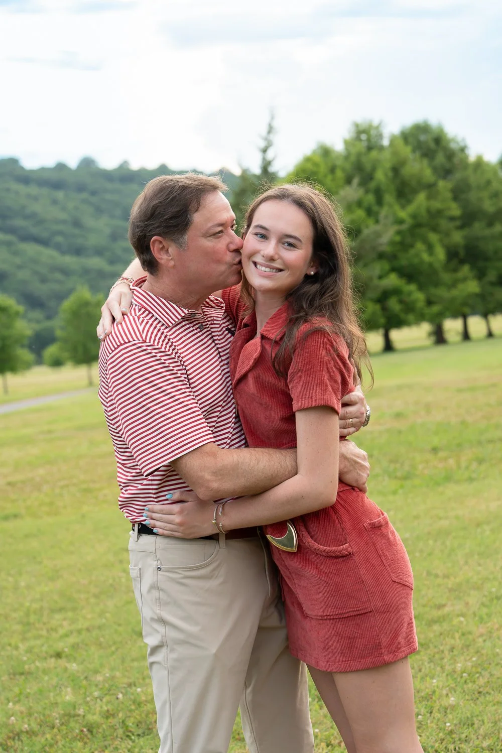 A man kissing a young woman on the cheek in a grassy park with trees and hills in the background.