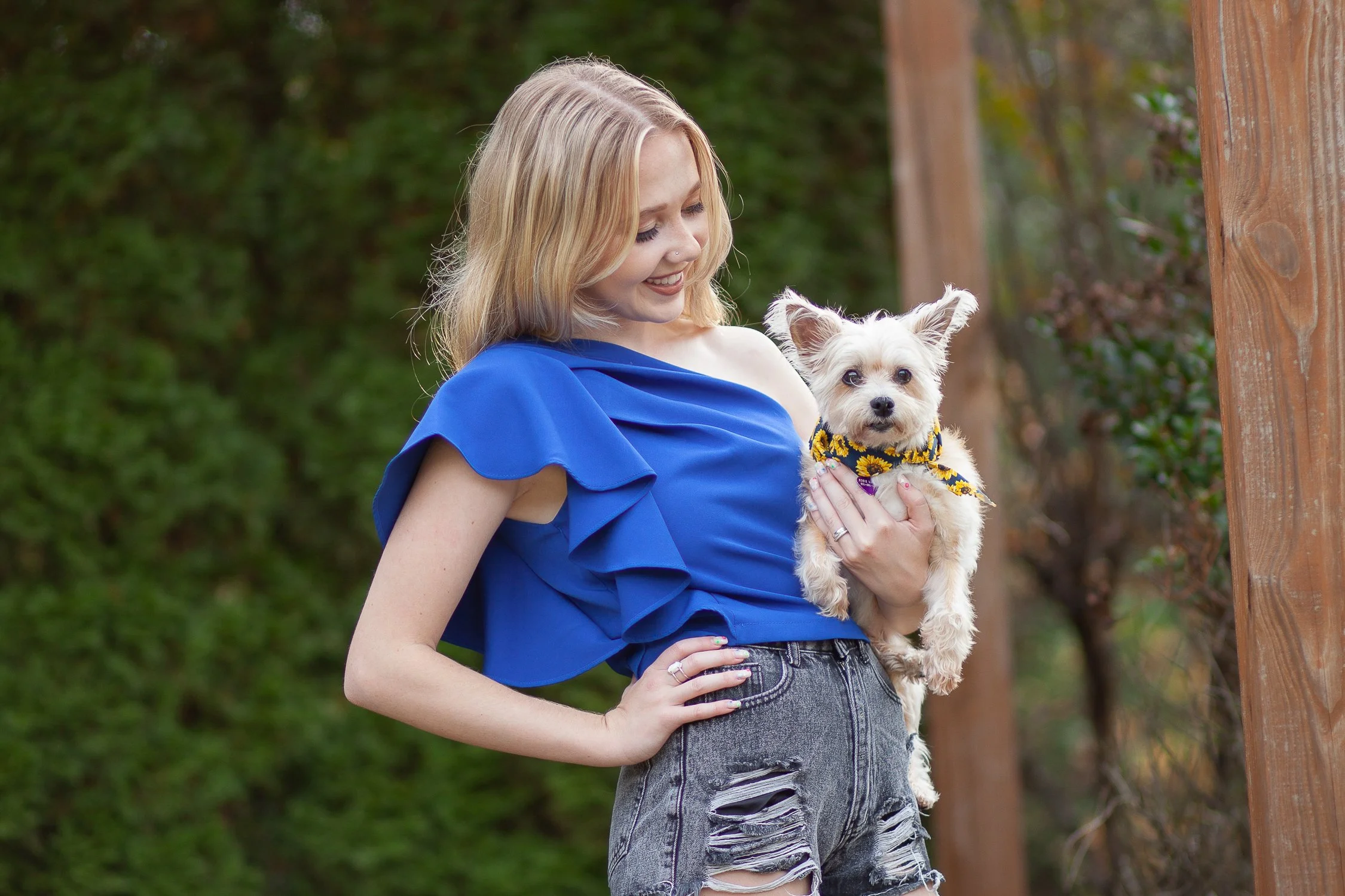 A young woman with blonde hair wearing a blue ruffled top and distressed denim shorts holding a small white dog with a sunflower bandana, standing outdoors with trees in the background.