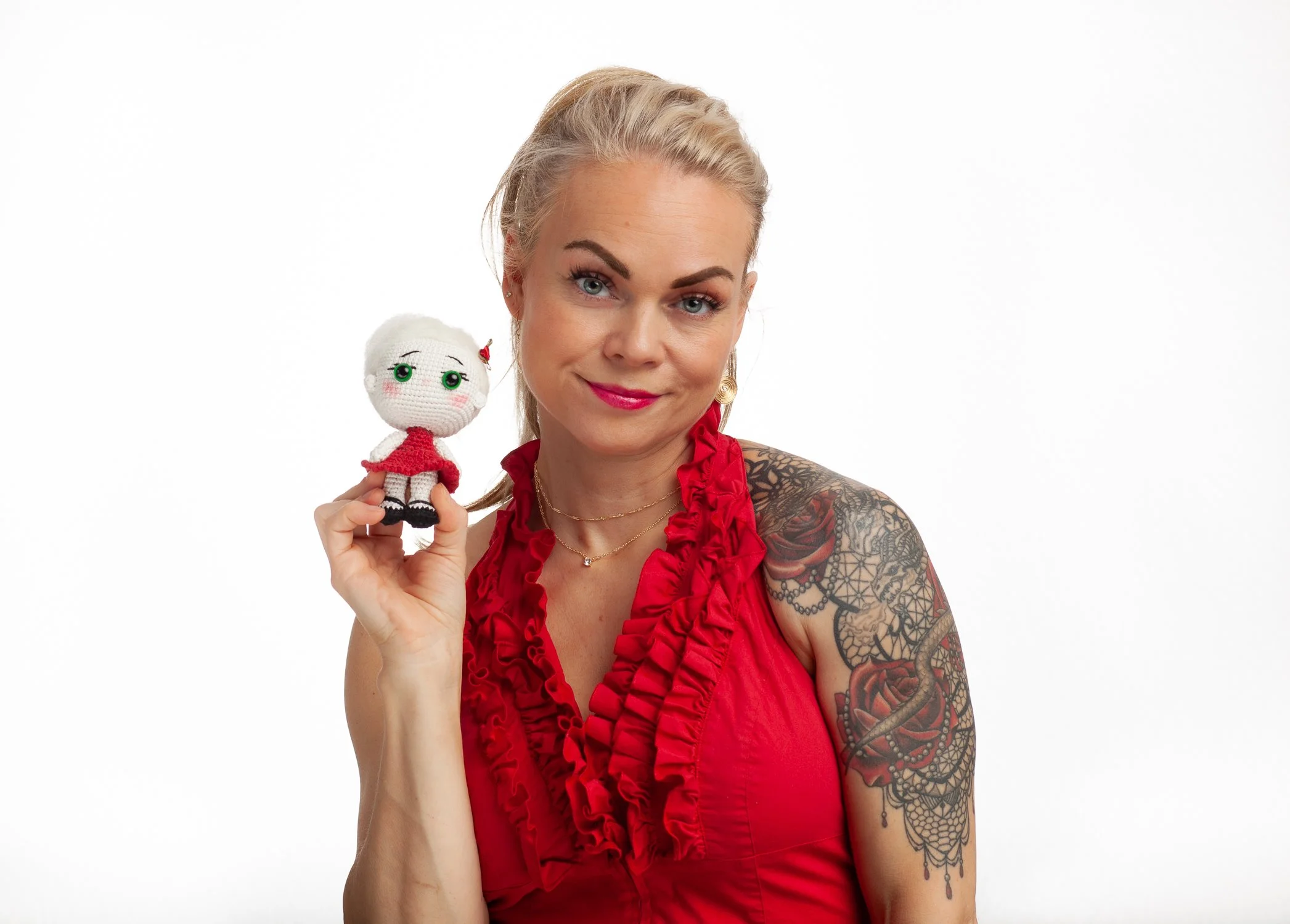 A woman with blonde hair, blue eyes, and tattoos on her left arm, wearing a red ruffled sleeveless top, holding a small crocheted doll with green eyes and red dress, smiling against a plain white background.