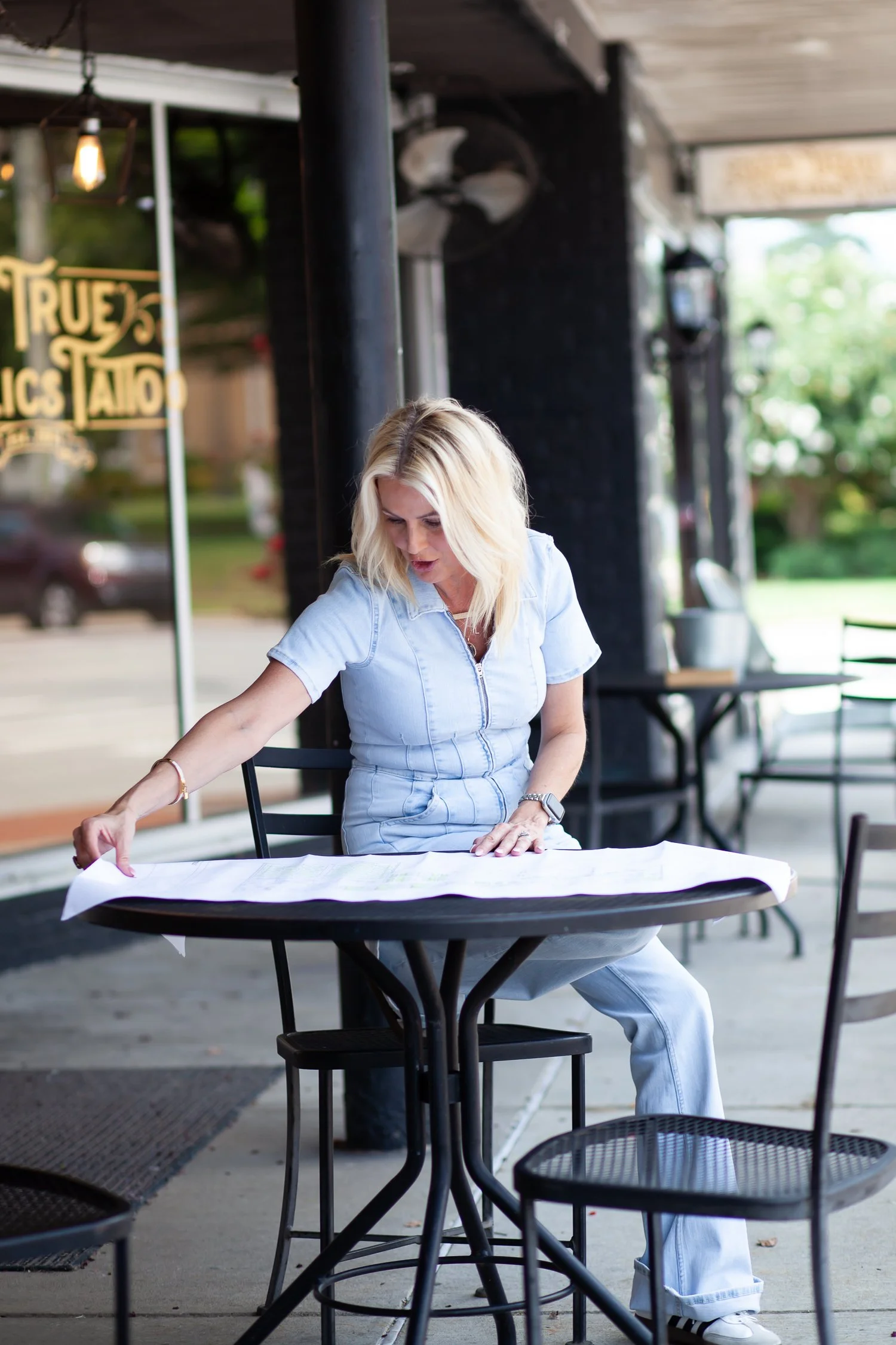 A woman with blonde hair, wearing a light denim outfit, sitting at an outdoor table, analyzing large papers or maps.