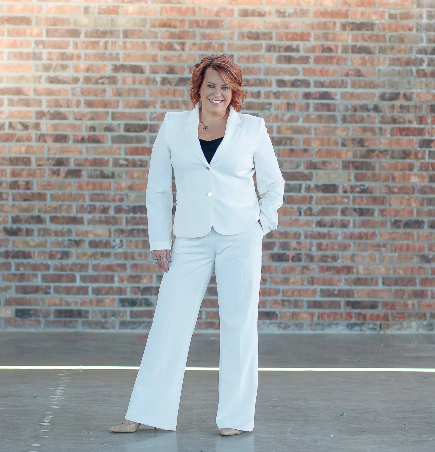 A woman with short red hair wearing a white pantsuit and beige heels, standing in front of a brick wall, smiling with her hands in her pockets.