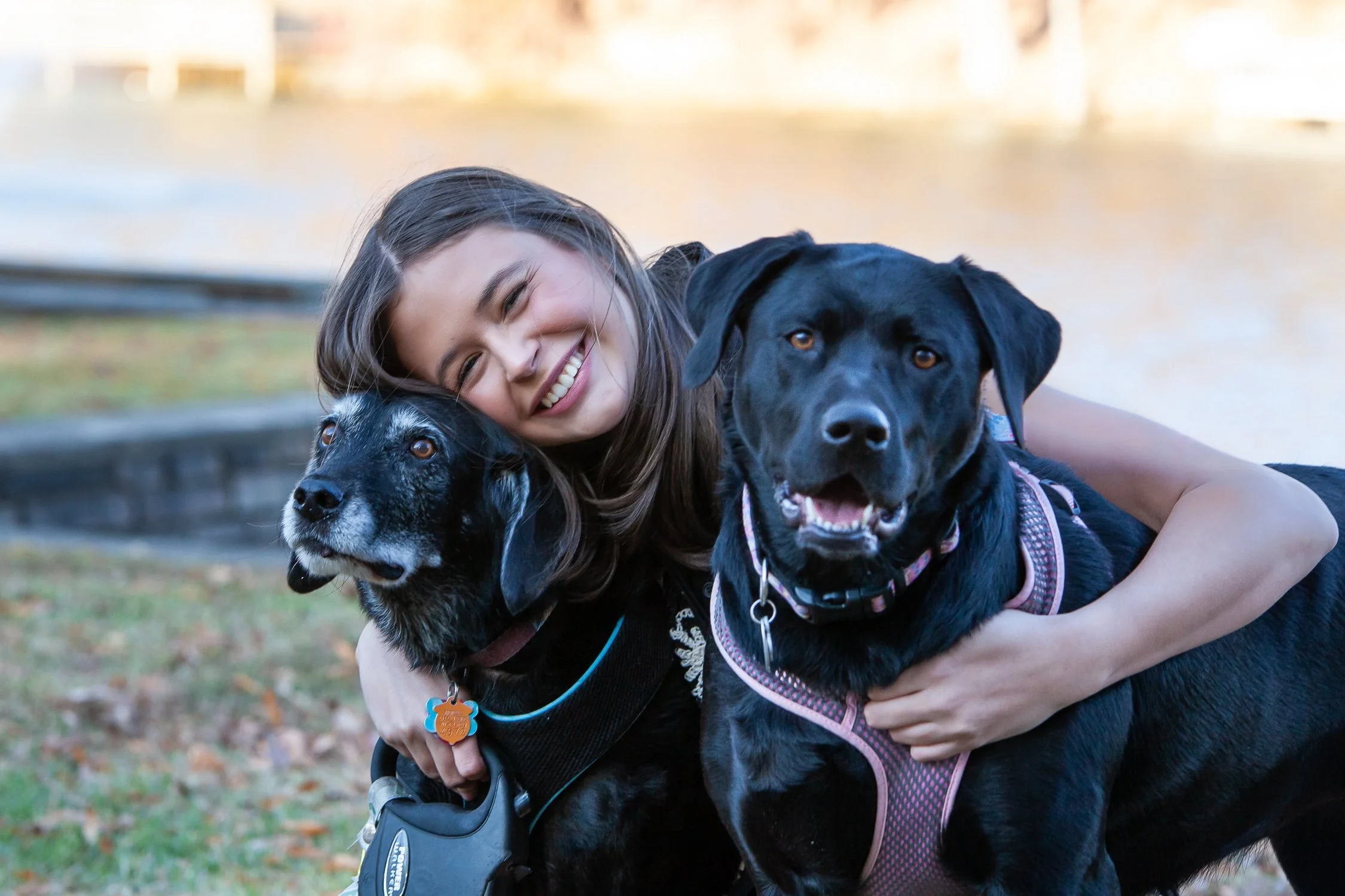 A young woman smiling and hugging two large dogs outdoors near a body of water.