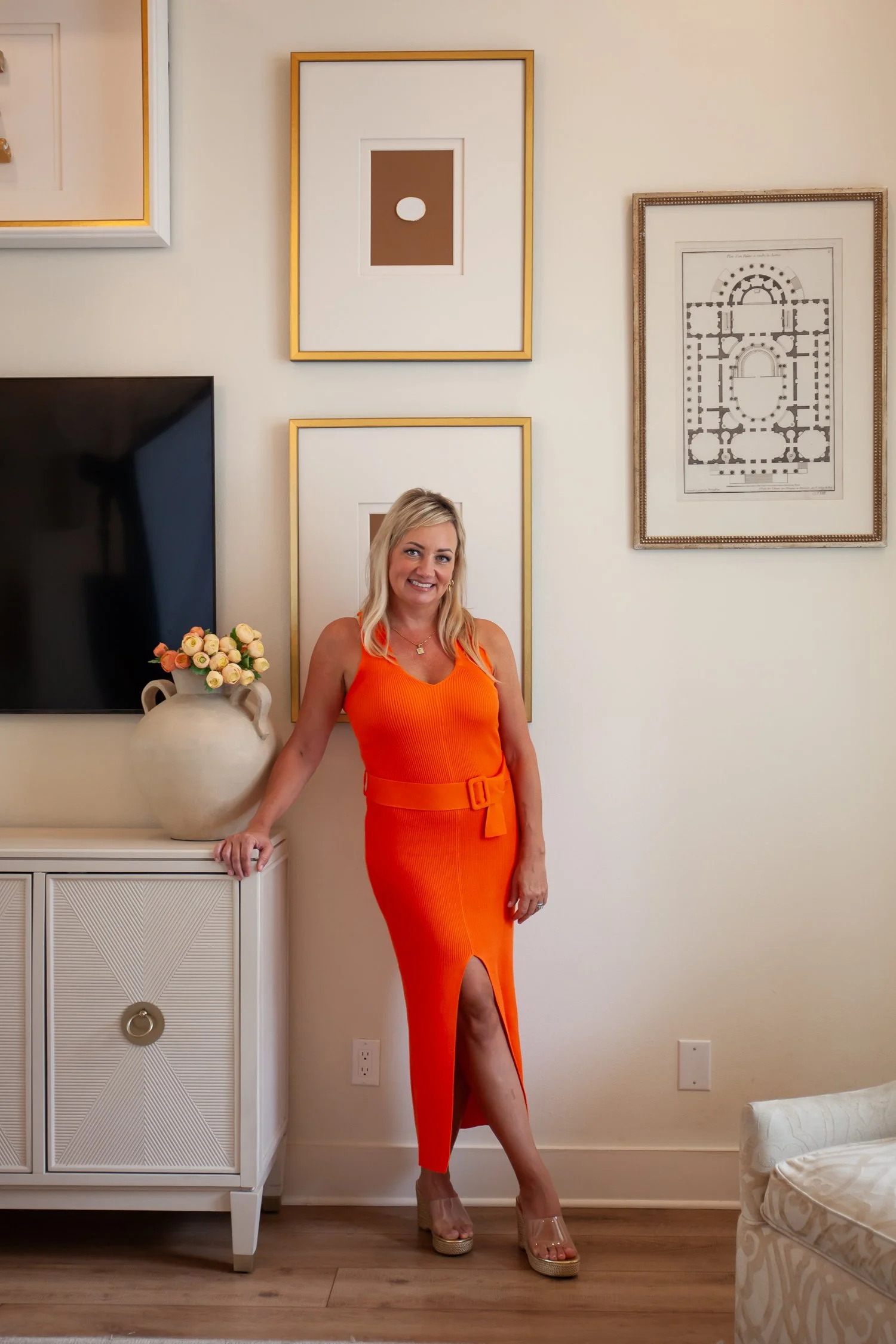 A woman with blonde hair wearing an orange dress with a slit, standing next to a white cabinet with a large flower vase on it, in a room decorated with framed artwork on the wall.
