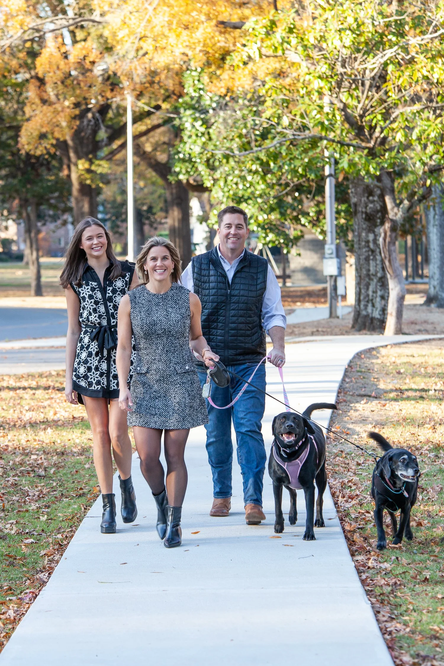 A family of three walking on a sidewalk in a park, with two black dogs on leashes. The background features trees with autumn foliage.