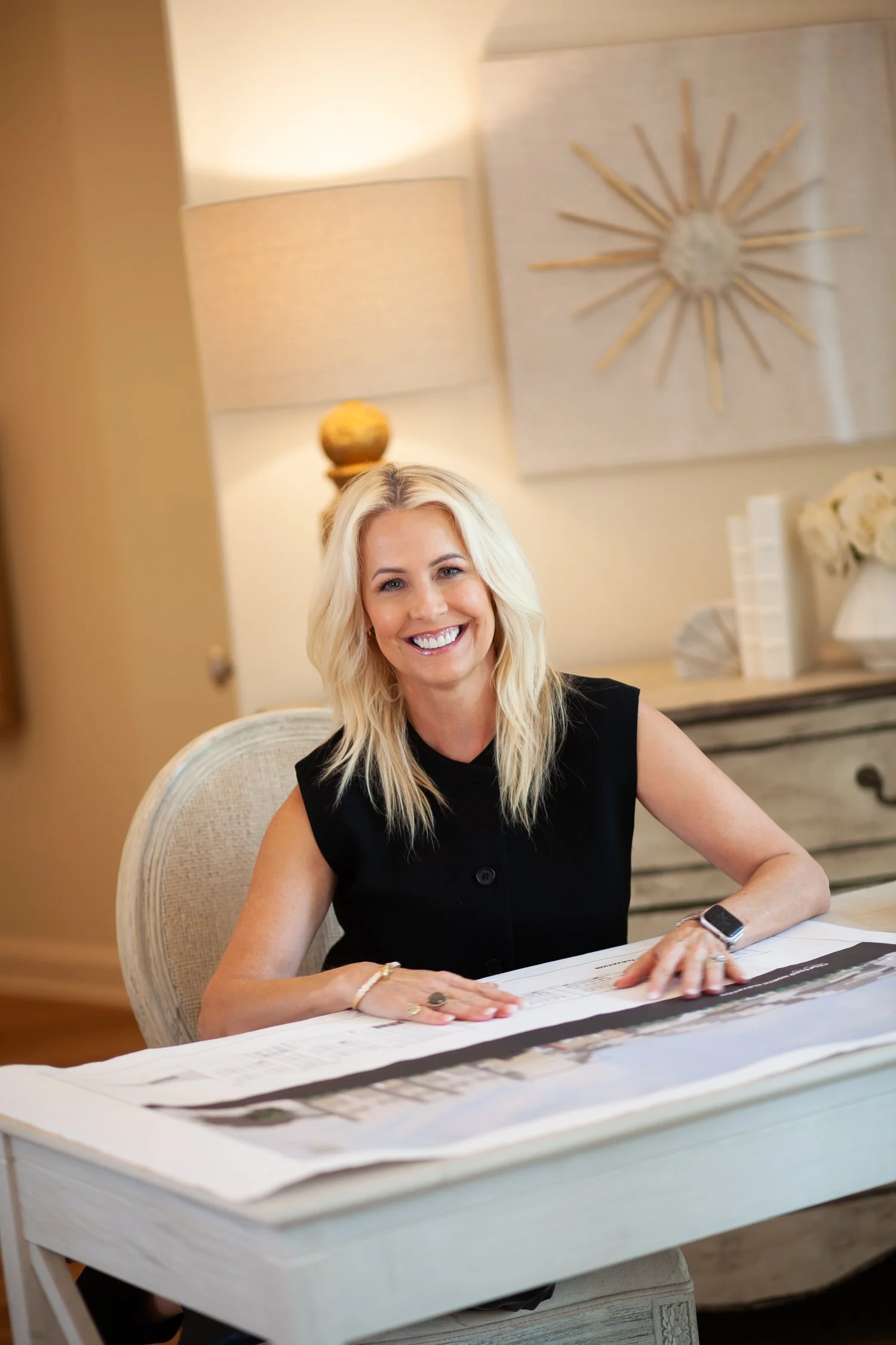 A smiling woman with blonde hair sitting at a white table with architectural blueprints, in a well-lit room decorated with a large wall art piece and a lamp.