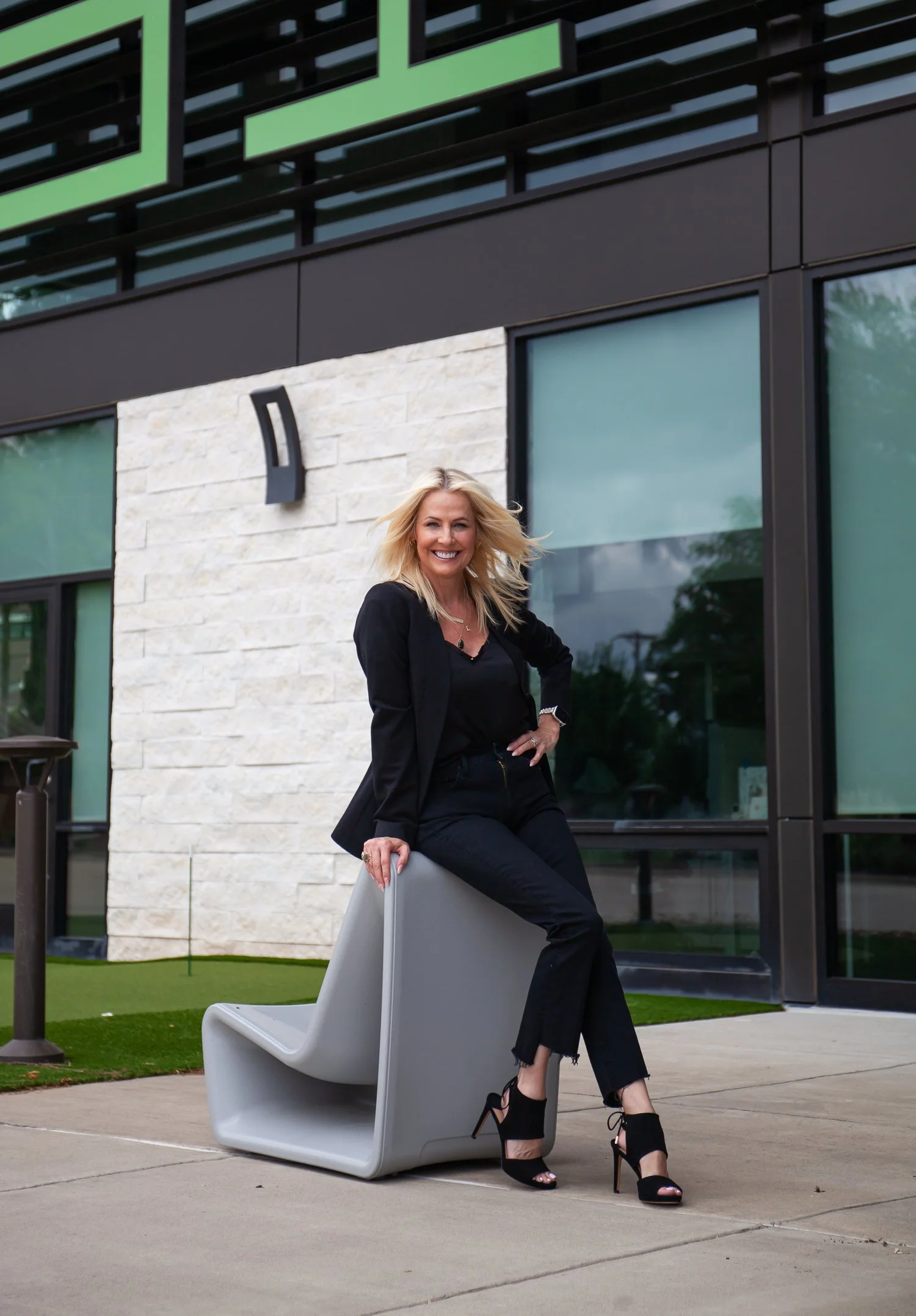 A woman with blonde hair, wearing an all-black outfit and high heels, sitting on a modern outdoor sculpture in front of a contemporary building with large windows and exterior lighting.