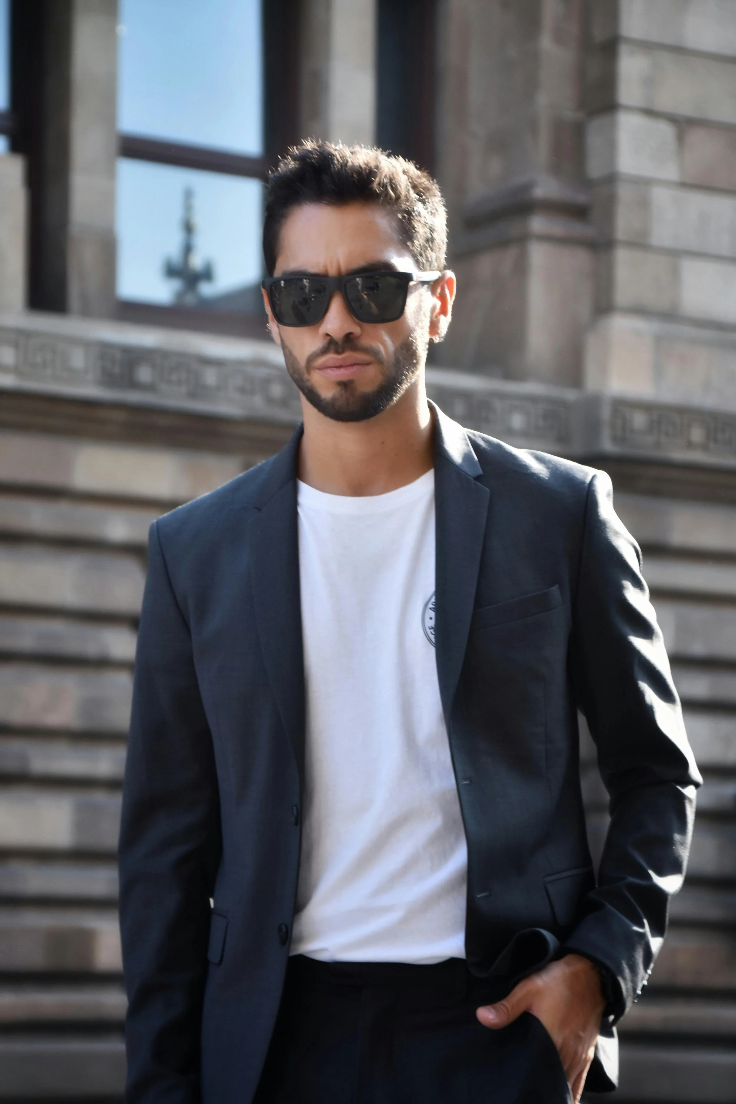 Joven con barba, gafas de sol negras, vestimenta formal con saco oscuro y camiseta blanca, en un entorno urbano con escaleras y edificio de piedra.