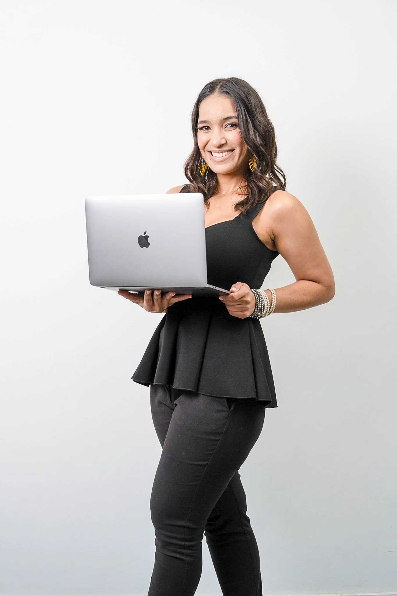 Mujer joven con vestido negro y joyas, sosteniendo una laptop plateada y sonriendo.