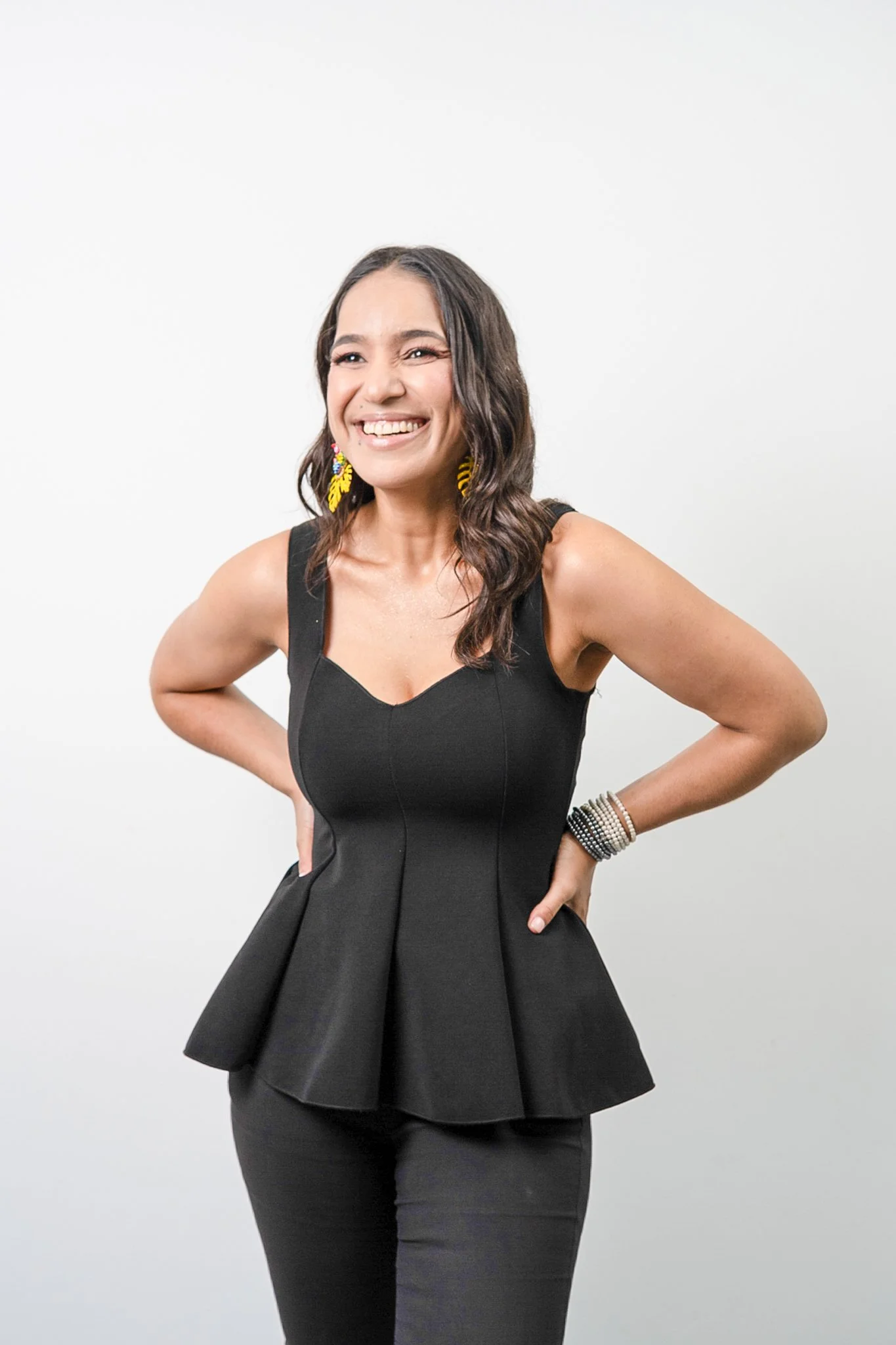 Mujer sonriendo, con vestido negro con falda de volantes, cabello rizado, aretes amarillos y pulseras en la muñeca derecha, posando con las manos en la cintura.