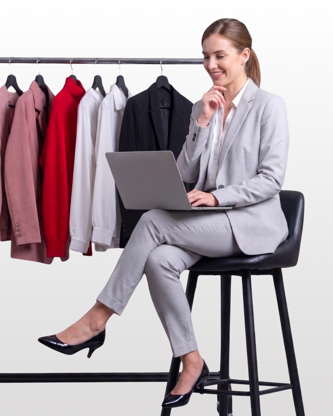 Una mujer con traje gris sentada en un taburete negro, usando una laptop, frente a una perchera con varias prendas de ropa. Son blusas y chaquetas en tonos rosado, rojo, blanco y negro.