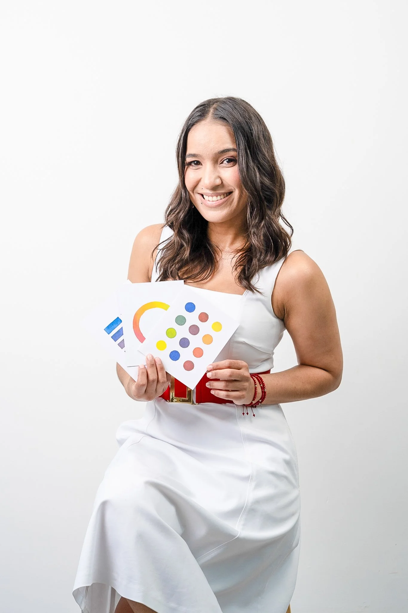 Mujer con vestido blanco y cinturón rojo sosteniendo papel con patrones de colores en fondo blanco.